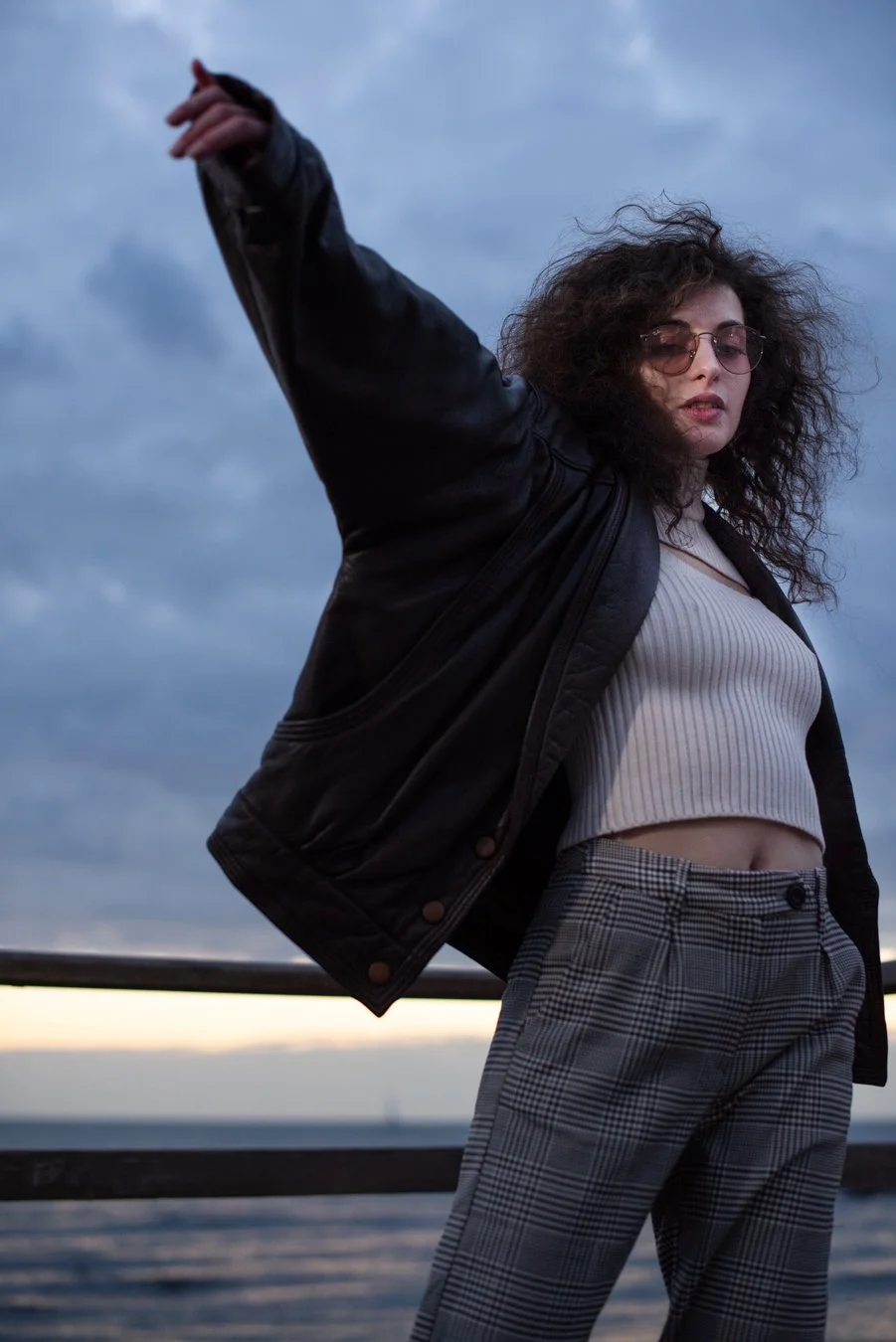 Donna con capelli ricci e occhiali da sole, indossa una giacca di pelle nera, una maglietta a righe corta e pantaloni a quadri, si trova vicino all'acqua con un cielo nuvoloso sullo sfondo.