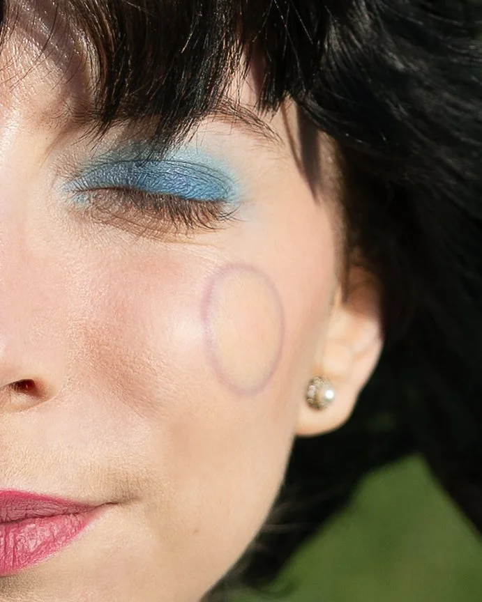 Parte del volto di una donna con trucco occhi blu, orecchino a perno e area circolare disegnata sulla guancia.