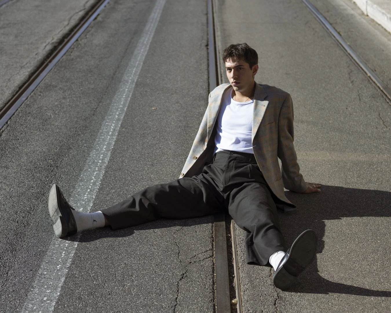 Un giovane uomo con giacca a scacchi, maglietta bianca, pantaloni neri e scarpe nere, è seduto a terra sulla strada con le gambe divaricate, tra le rotaie di un tram, sotto la luce del sole.