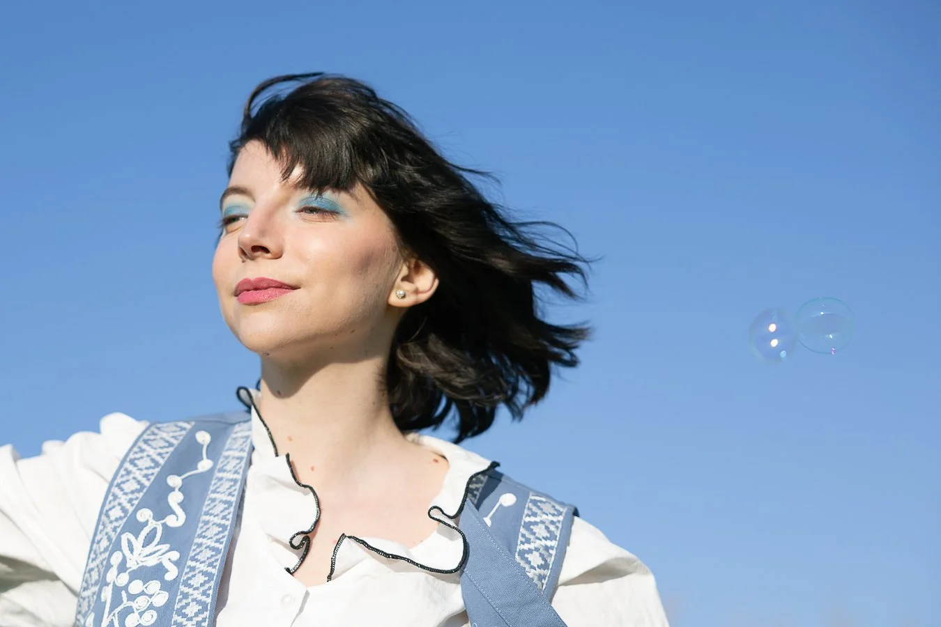 Una donna con i capelli corti neri che sorride con gli occhi chiusi, indossa un vestito bianco con dettagli blu e una borsa blu, si trova all'aperto sotto un cielo azzurro con bolle di sapone che volano.