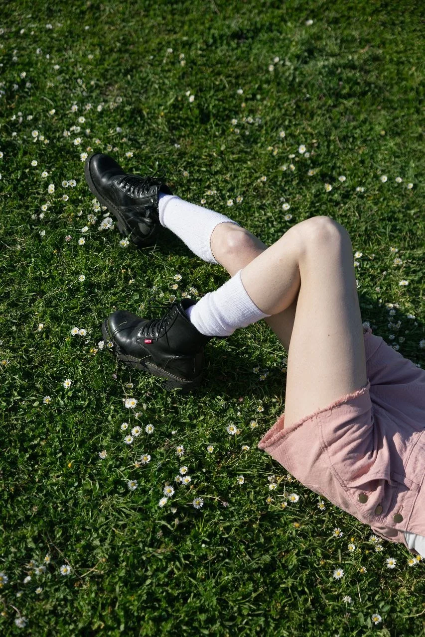 Gambe di una persona sdraiata sull'erba con scarpe nere, calzini bianchi, pantaloncini rosa e un campo di fiori bianchi e verdi