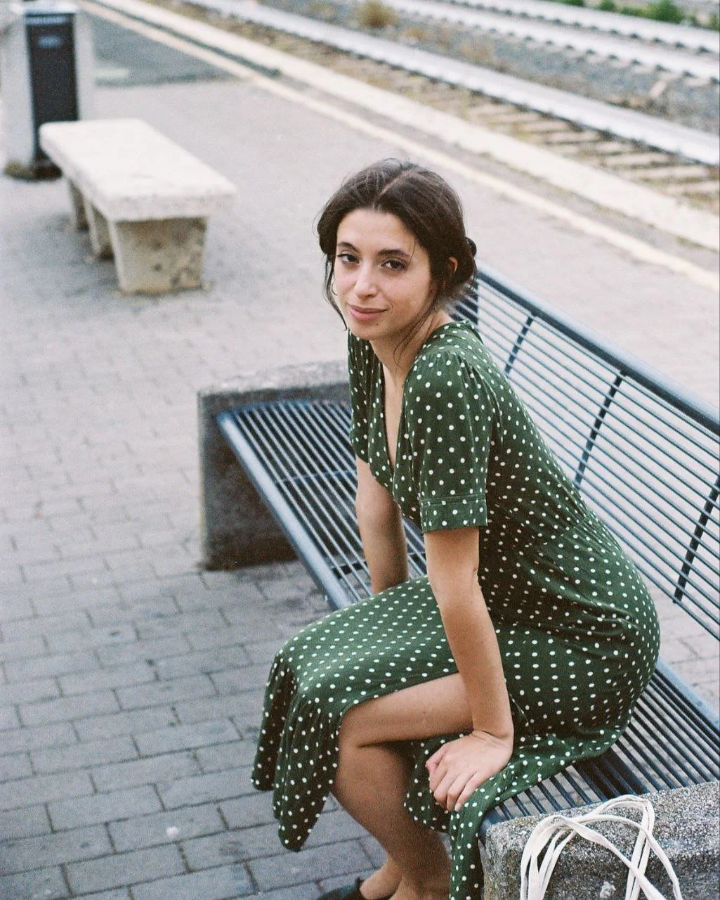 Una giovane donna seduta su una panchina in una stazione ferroviaria, indossa un vestito verde a pois bianchi, con i capelli raccolti e un sorriso leggero.