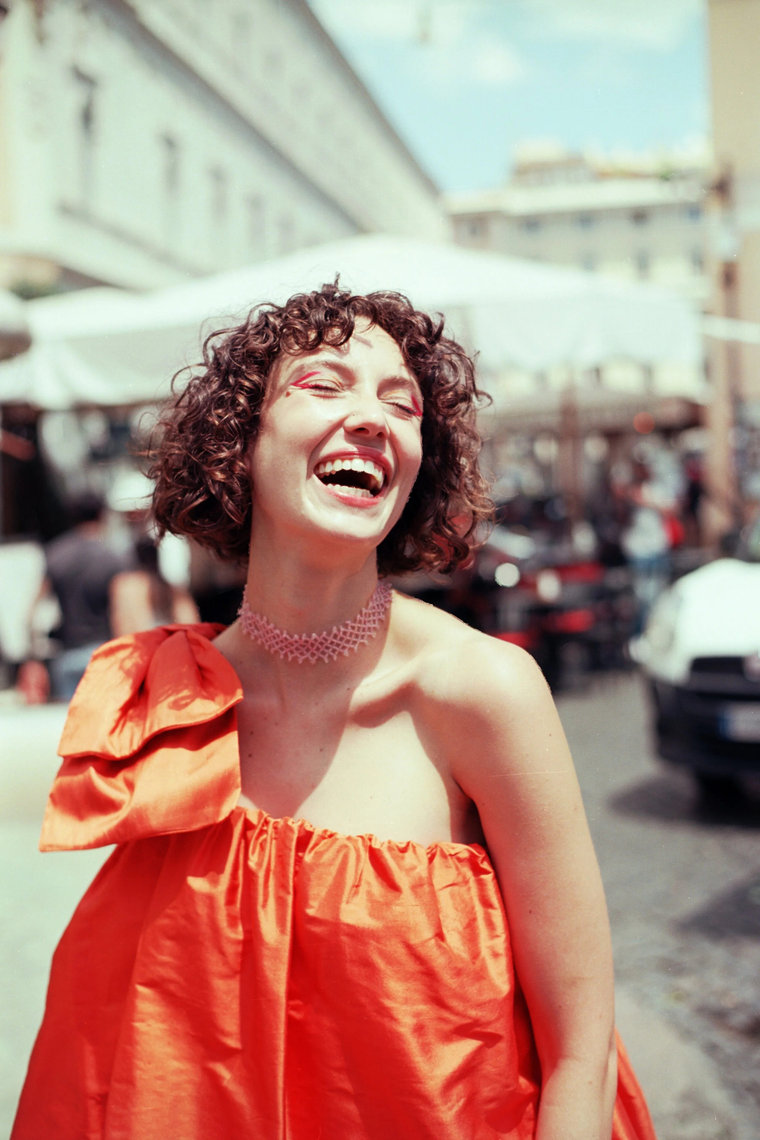 Una donna con capelli ricci, sorridente, indossa un vestito arancione con una spalla scoperta e un collanina rosa, sullo sfondo si vedono edifici e ombrelloni in un ambiente esterno soleggiato.