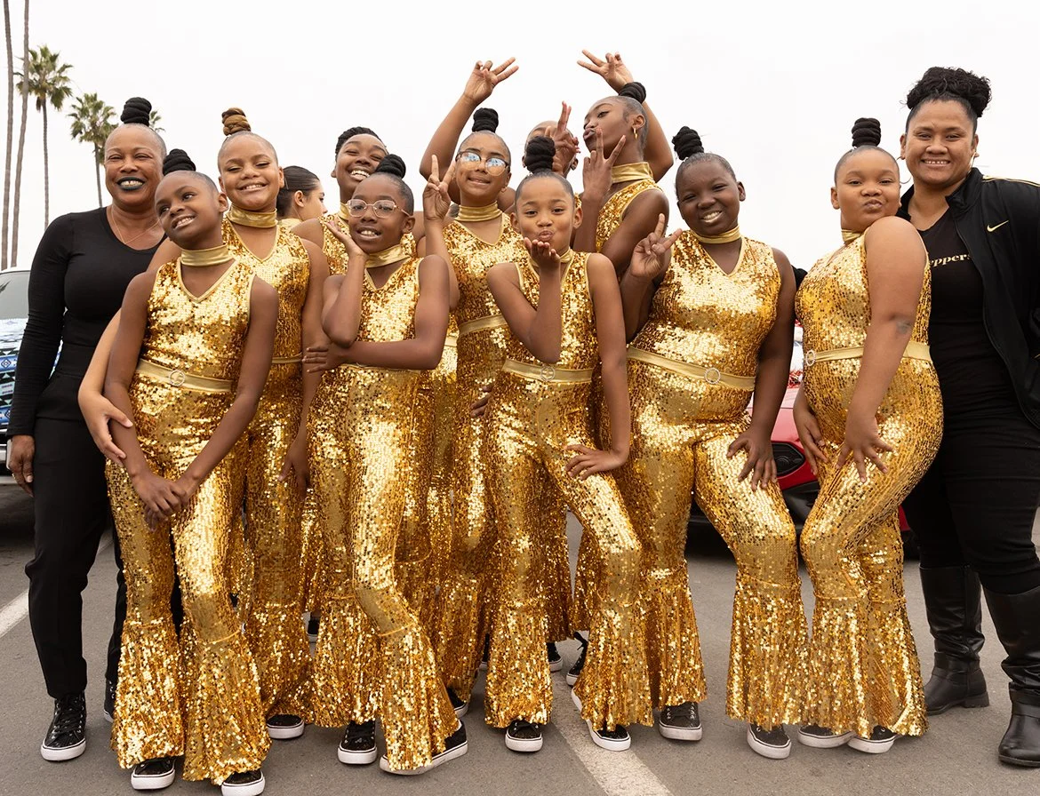 A group of young girls and two adult women posing outdoors, all dressed in matching gold sequin outfits, with palm trees in the background at a massive parade event.