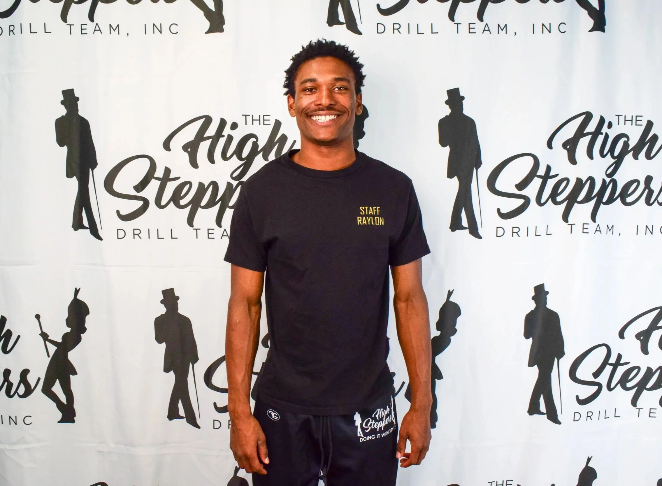 A member of the High Steppers team staff stands with curly hair wearing a black t-shirt and black shorts in front of a backdrop with silhouettes of leaders and the logo for The High Steppers Drill Team, Inc.