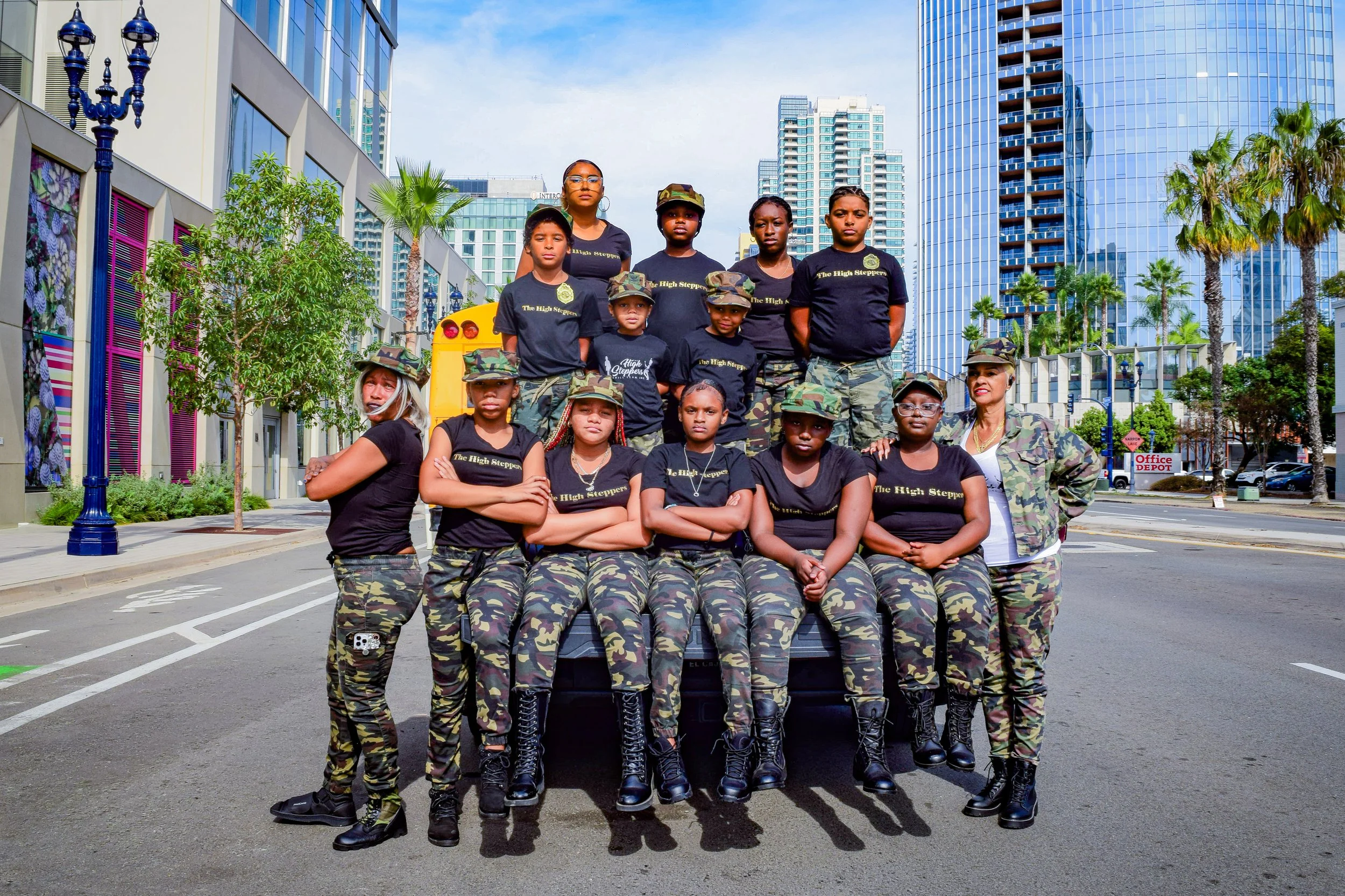 The High Steppers dressed in camouflage clothing, standing and sitting in front of a yellow vehicle on a city street with downtown buildings and palm trees in the background.