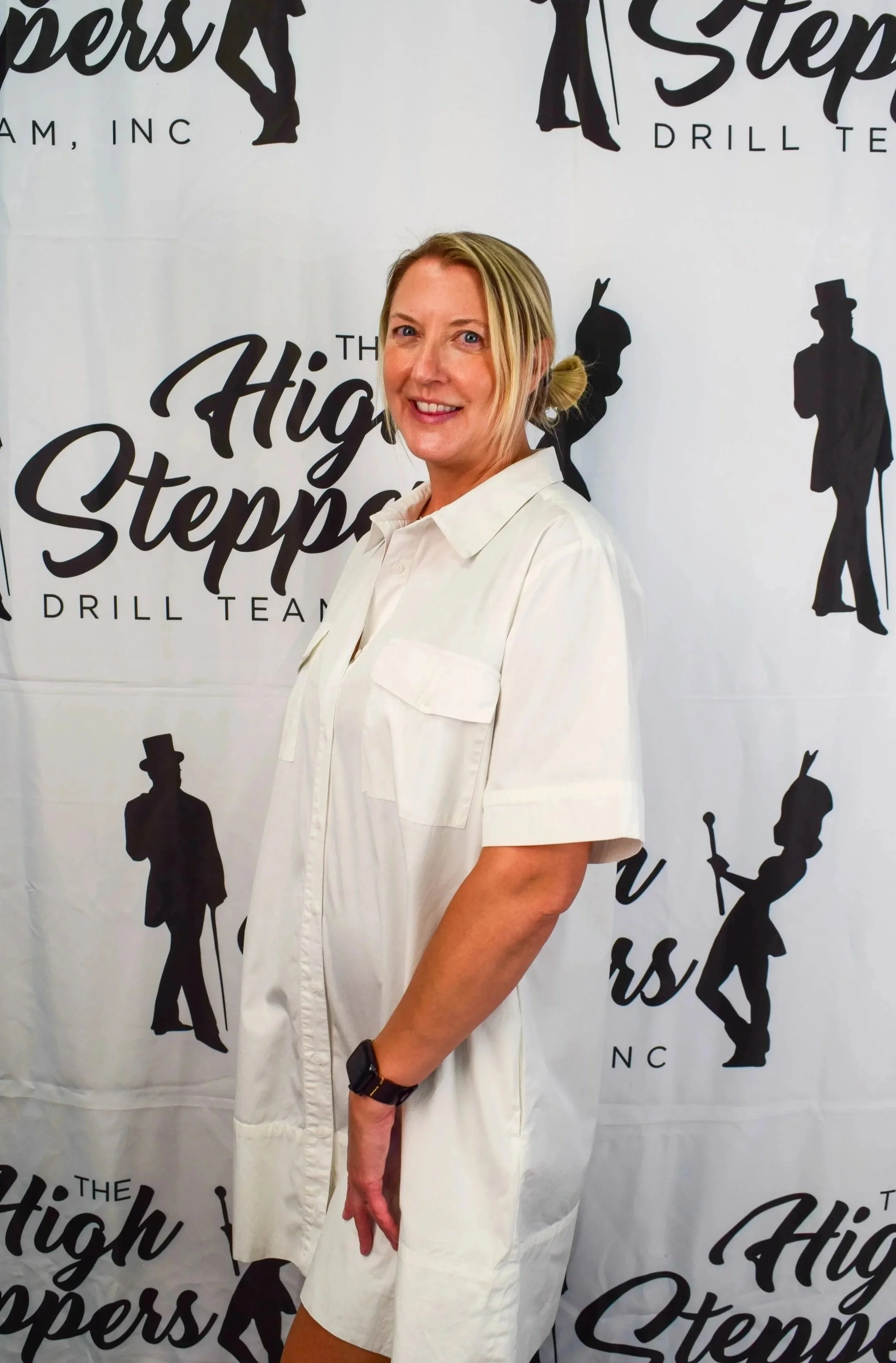 A member of the High Steppers team staff stands wearing a white dress and a black wristwatch, standing in front of a backdrop with the logo of The High Steppers drill team featuring silhouettes of leaders.
