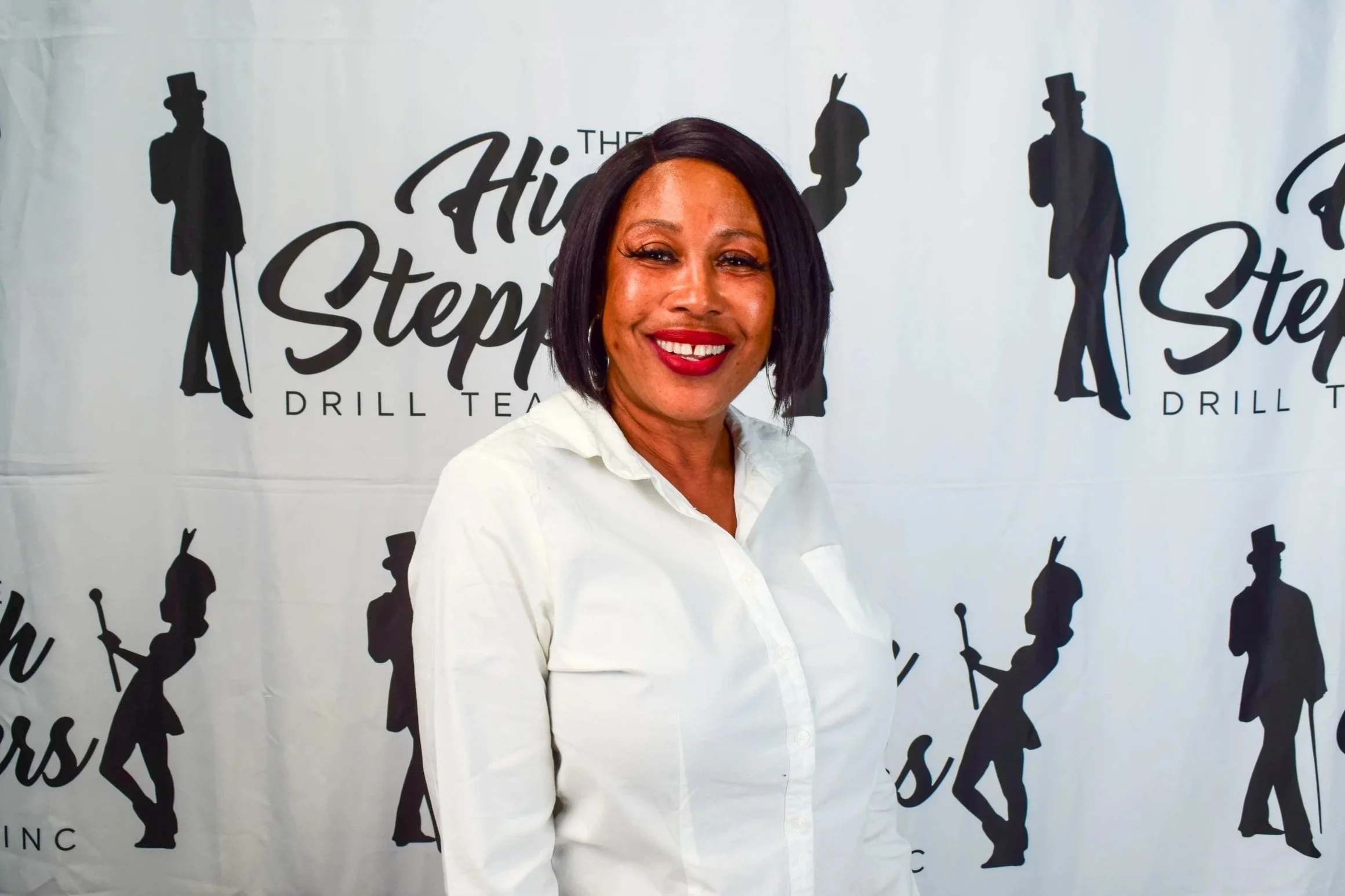 The Founder and CEO of the High Steppers Drill Team Inc. Stands with shoulder-length black hair smiling at the camera, wearing a white shirt, in front of a backdrop with black silhouettes of the high steppers logo.