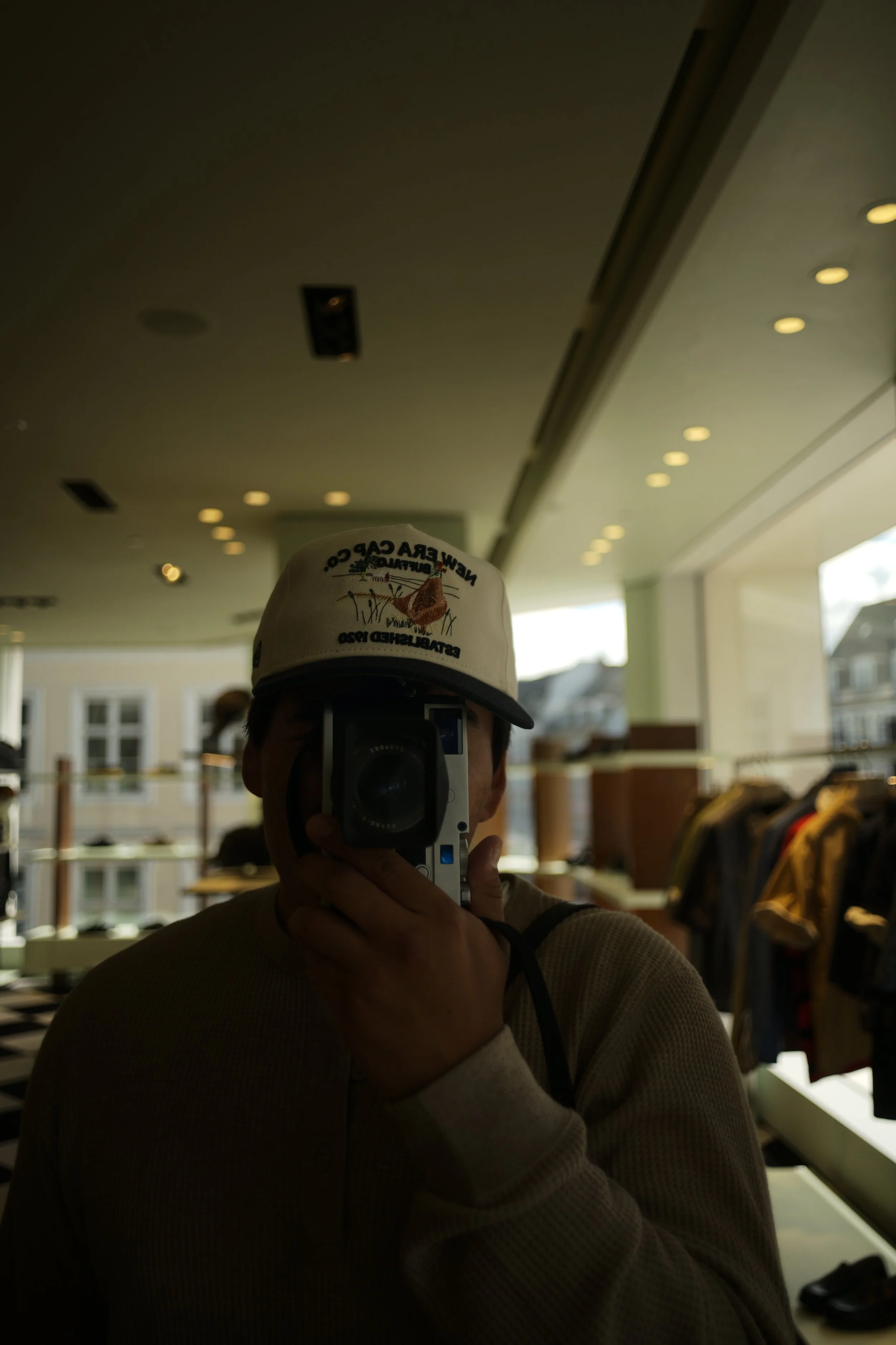 Person taking a photo of their reflection in a mirror in a clothing store, wearing a baseball cap and a beige sweater.