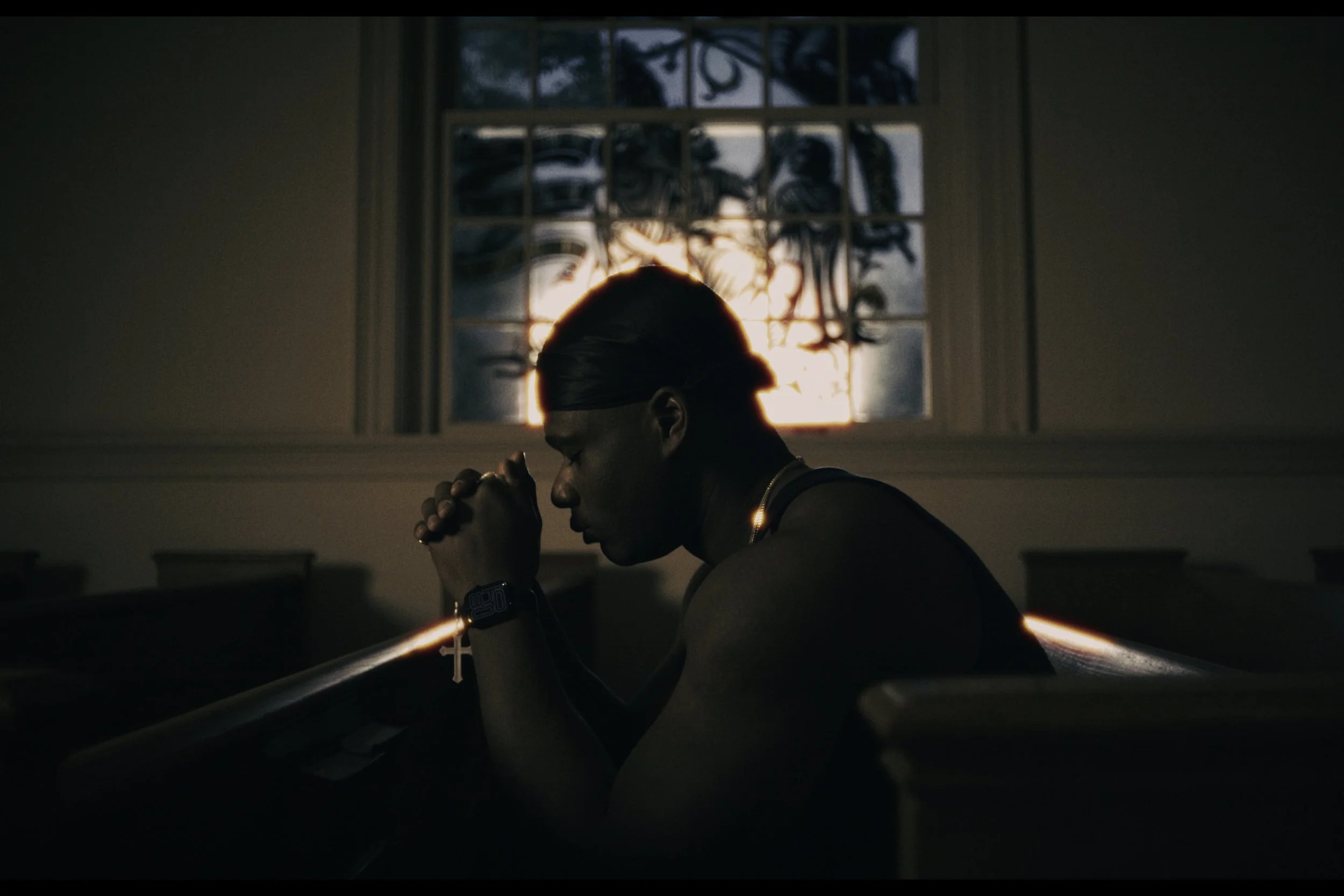 A person with short hair and a headband sits with their eyes closed and hands clasped in a prayer position in a dimly lit room, with sunlight shining through a window showing trees outside.