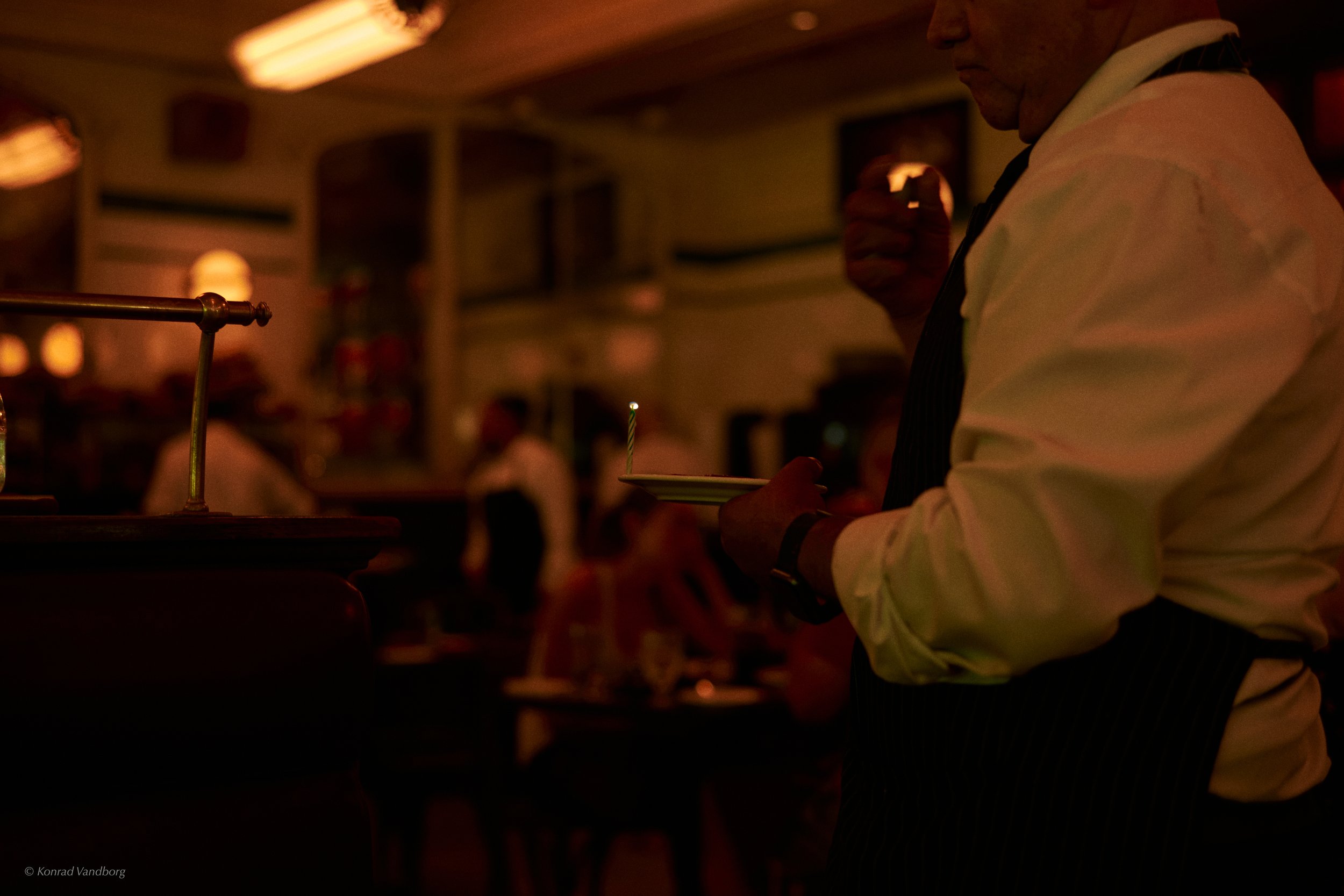 A person wearing a white shirt and dark apron holding a small plate with a lit birthday candle in a dimly lit restaurant or bar.