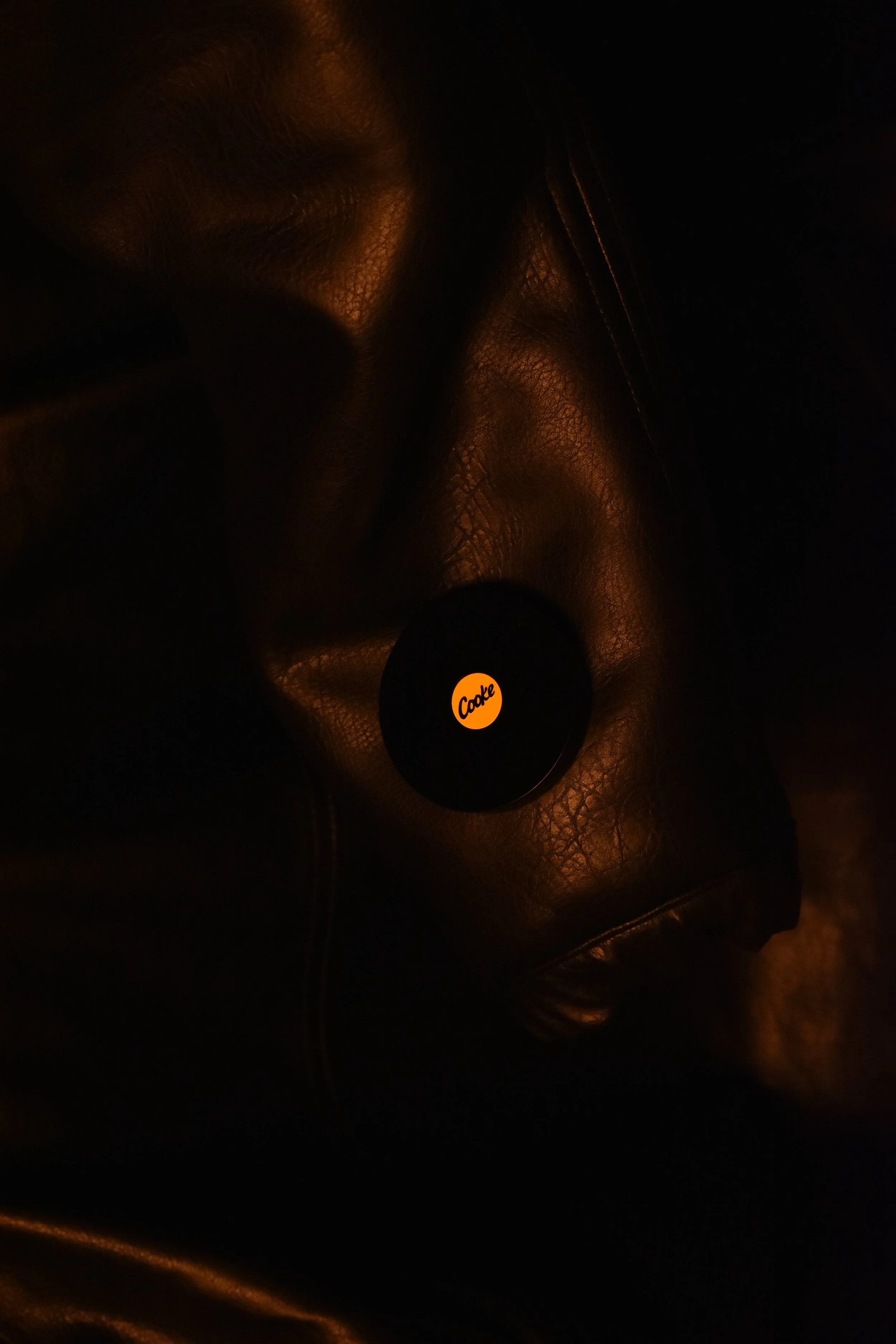 A dark image showing a shiny, textured black leather surface with a small circular object at the center with the word 'Cooke' in orange. The lighting reflects off the leather, creating a warm glow.