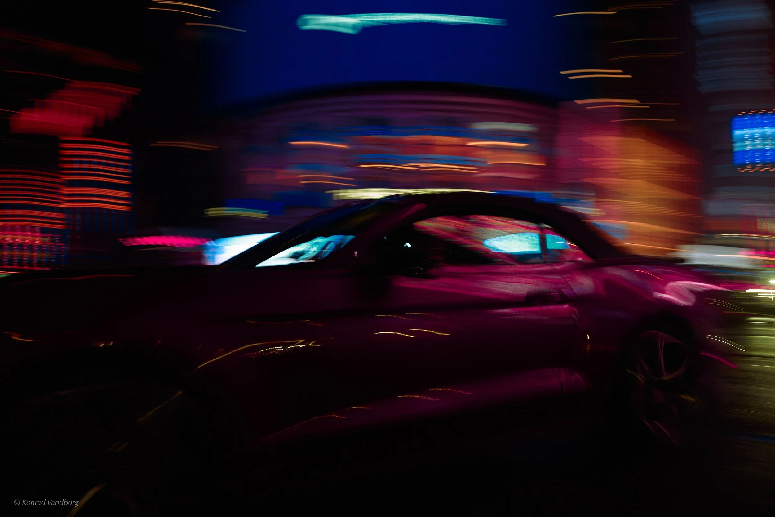 Long exposure photo of city lights at night with colorful streaks of red, blue, yellow, and pink, and a dark silhouette of a car in the foreground.