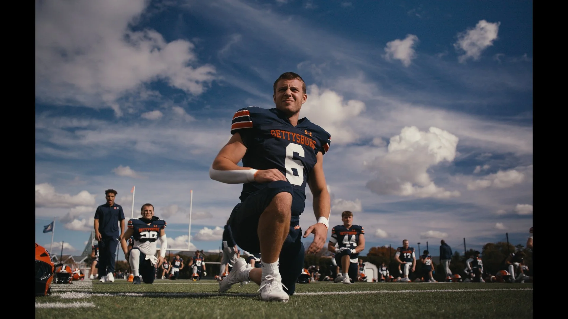 American football team in uniform warming up on the field before a game, with a player kneeling in the foreground and others standing in the background under a partly cloudy sky.