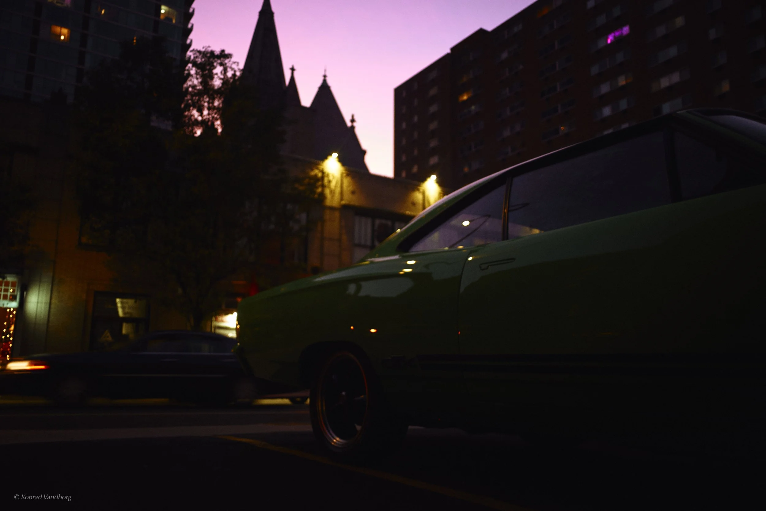 A green car is parked on a city street during dusk, with high-rise buildings and a church with tall steeples in the background, illuminated by streetlights and a purple-pink sky.
