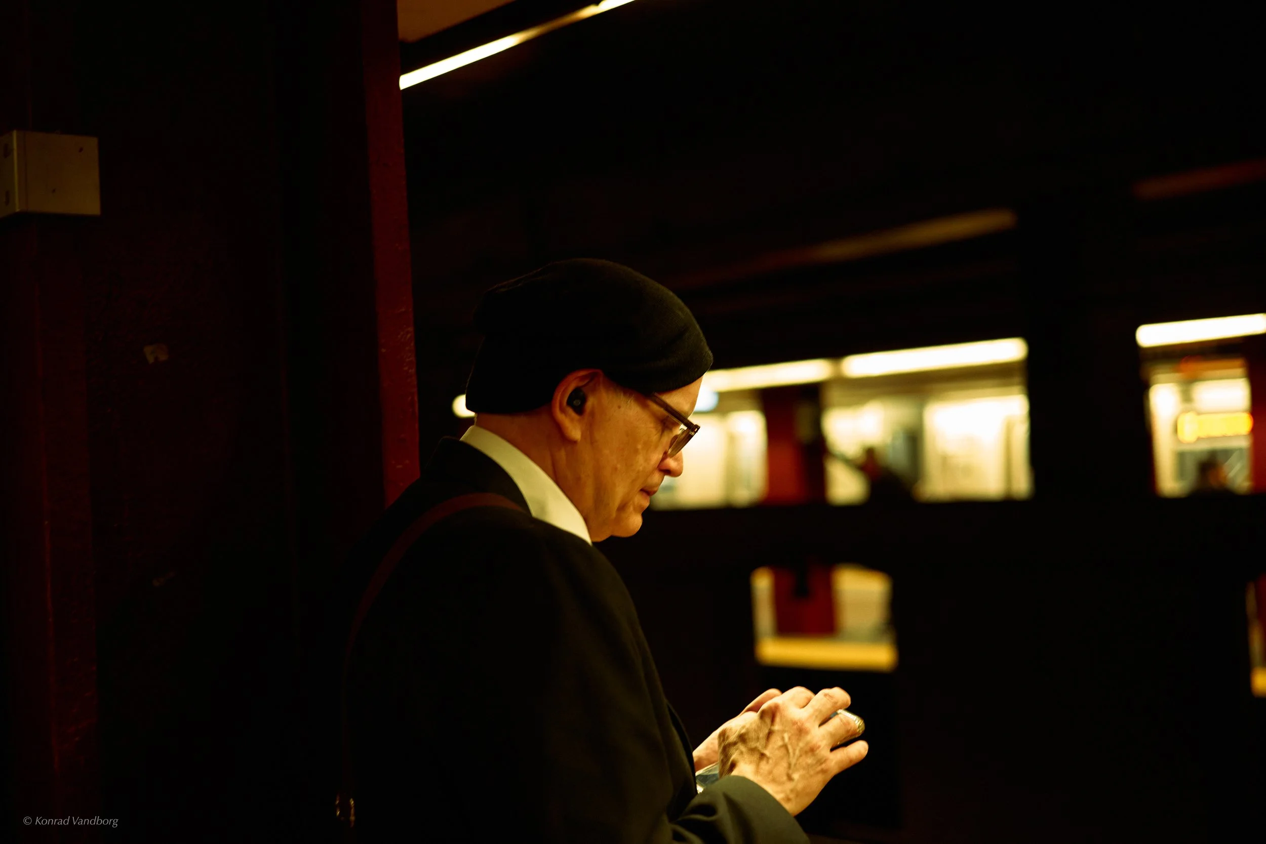 A man wearing a black beanie and glasses standing in a dimly lit space, looking at his phone with bright background lights.