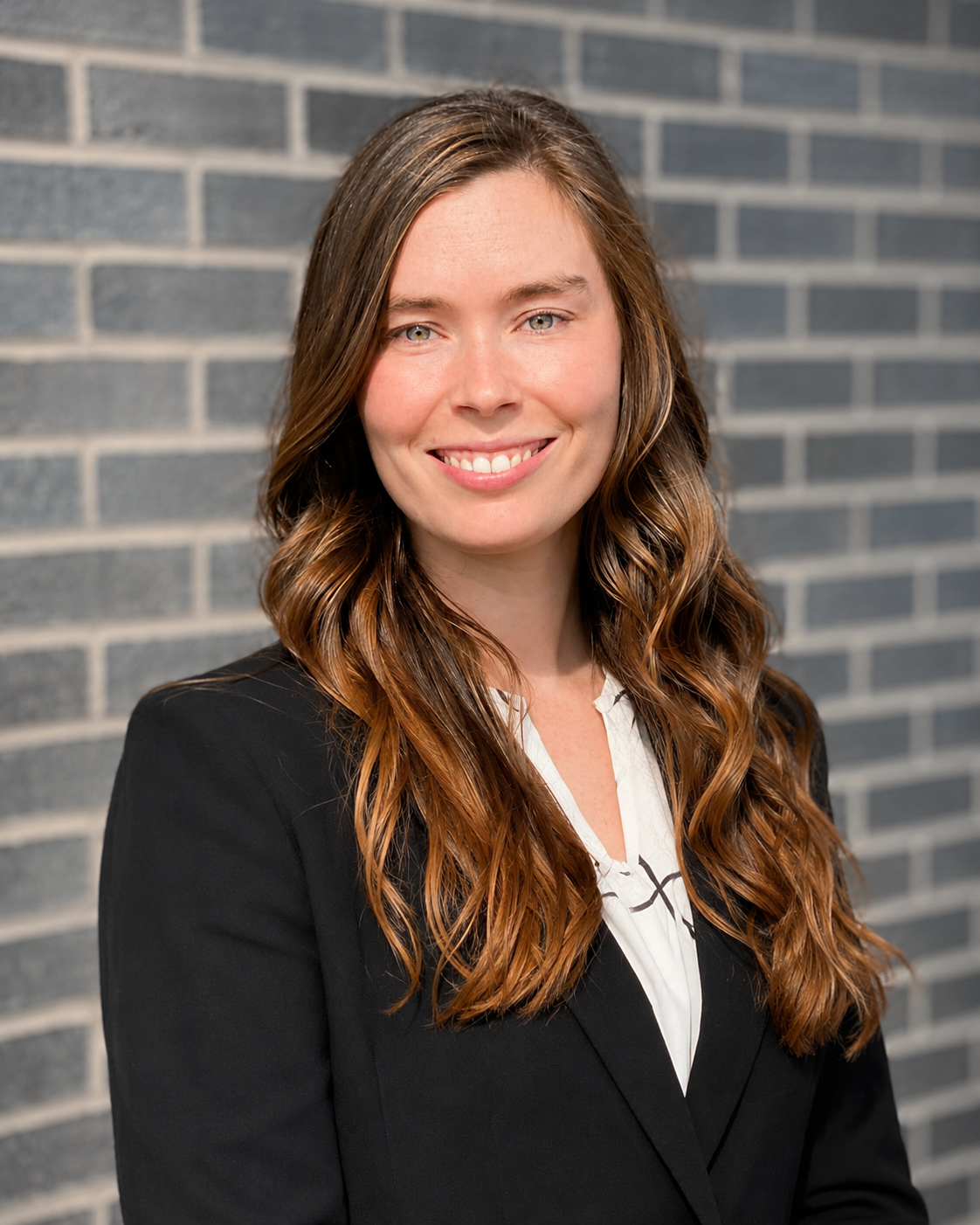 A professional woman with long wavy brown hair, wearing a black blazer and white blouse, smiling in front of a brick wall.