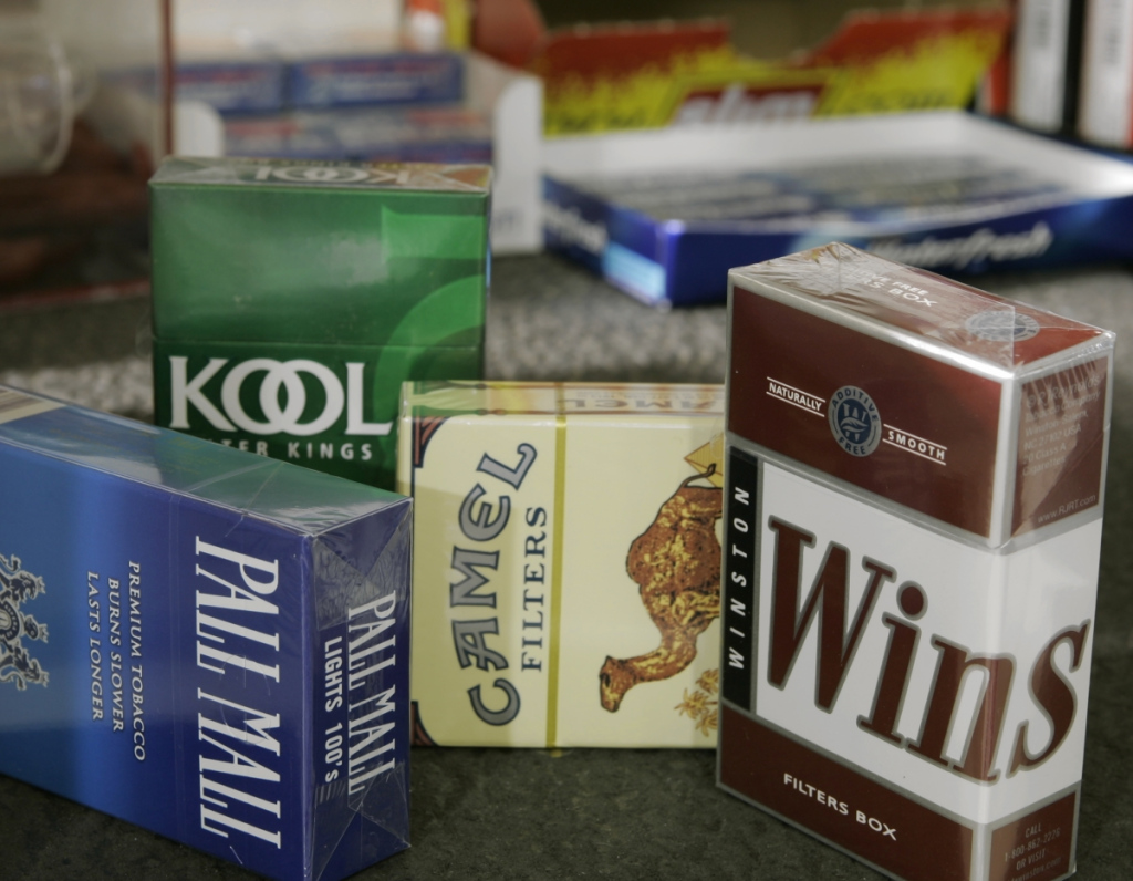 Various brands of cigarette packs on a surface, including Kool, Wins, and Pall Mall, with additional tobacco products blurred in the background.