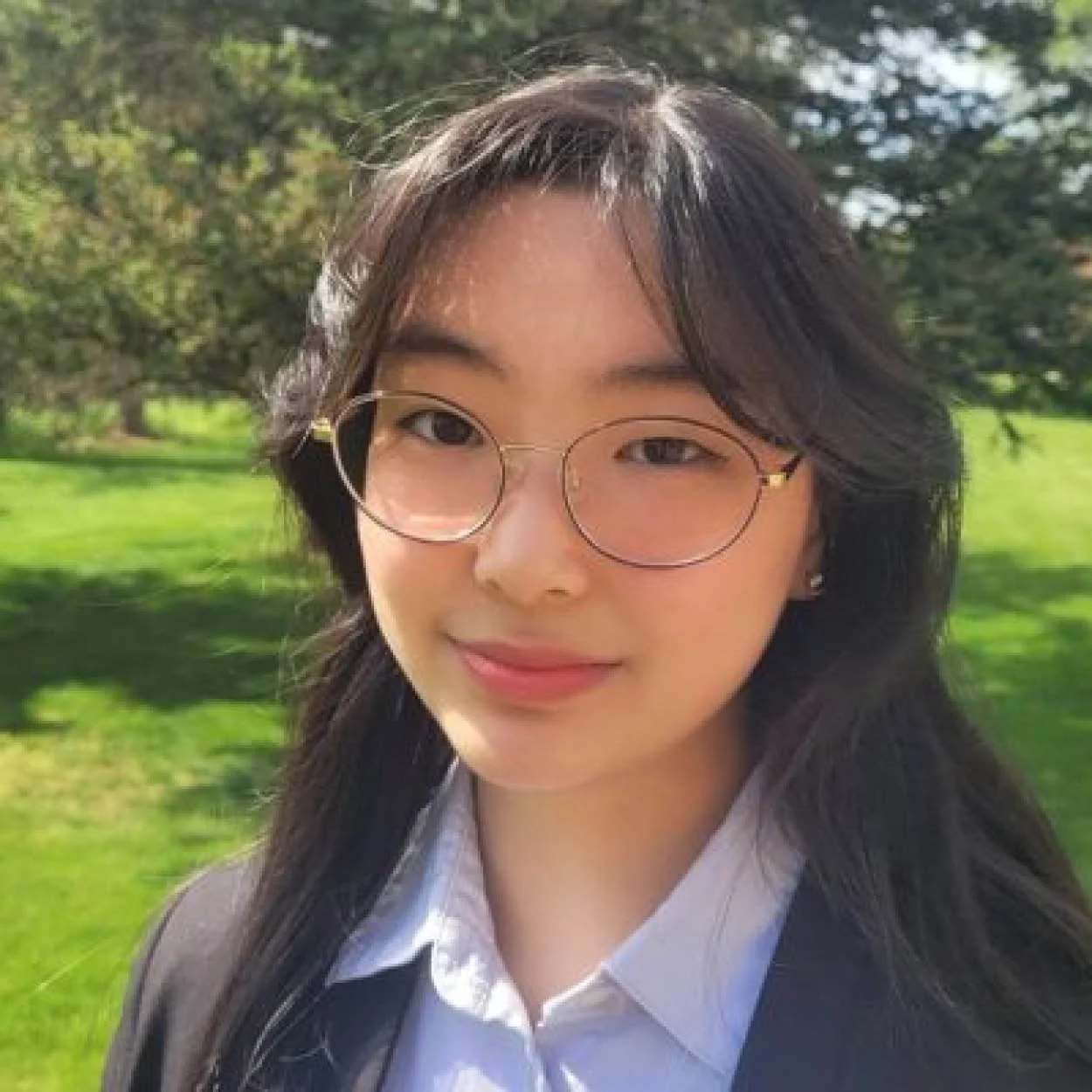 A young woman with long black hair wearing glasses, a white shirt, and a blazer, standing outdoors in a park with green grass and trees in the background.