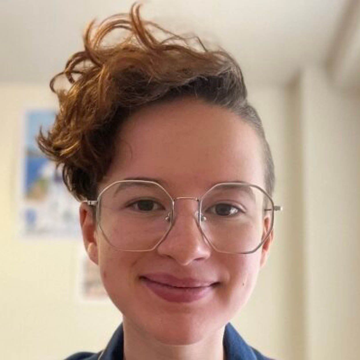 A person with short, curly, auburn hair and glasses smiling at the camera. The background shows a blurred wall with a picture or poster on it.