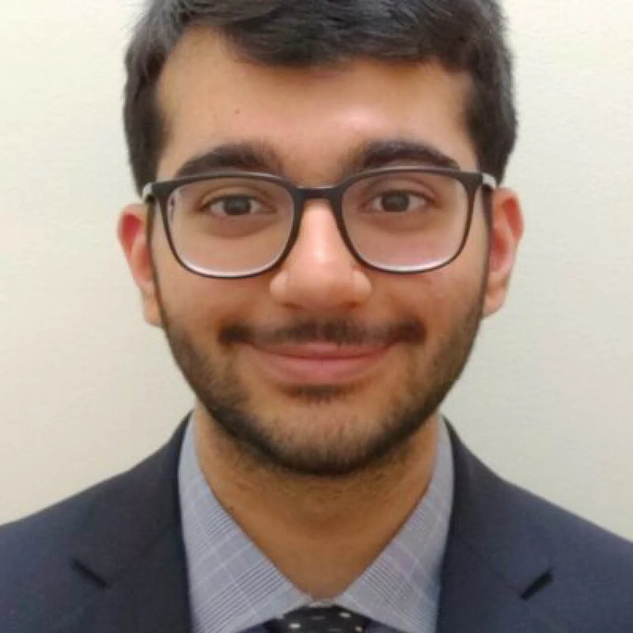 A young man with glasses, dark hair, and a beard wearing a suit and tie, smiling against a plain, light-colored background.