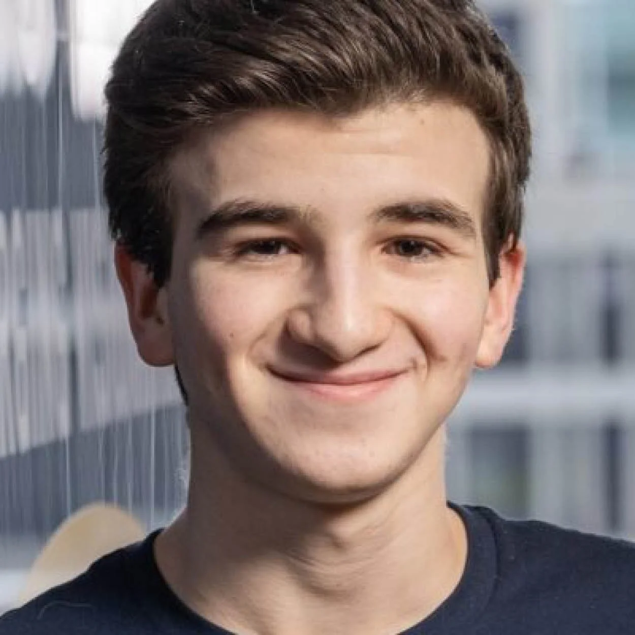 Close-up of a young man with brown hair and a smile, standing outdoors with a blurred cityscape in the background.