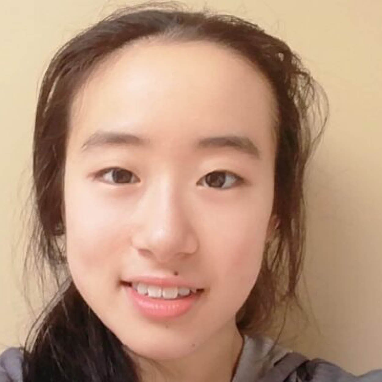 Close-up of a young woman's face with light skin, dark eyes, dark hair, smiling, against a beige background.