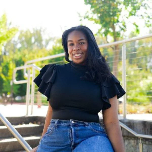 Young woman with long black hair smiling outdoors in front of railing and green trees, wearing a black ruffled sleeve top and blue jeans.
