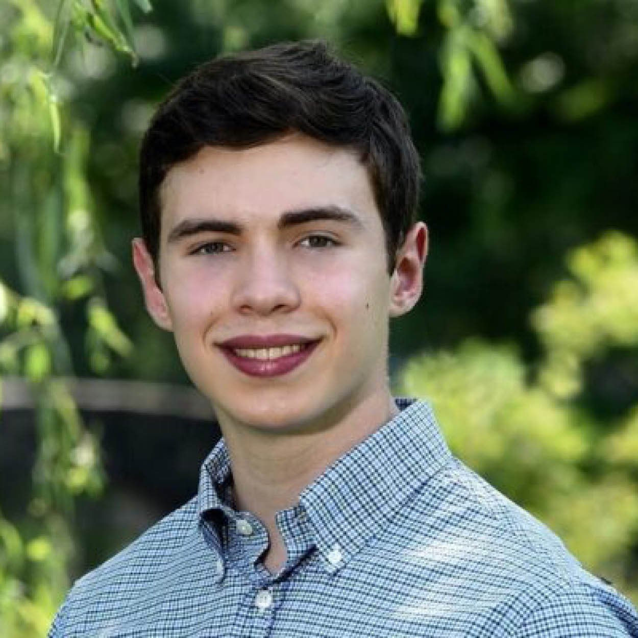 Headshot of a young man with short dark hair and light skin, wearing a checkered shirt, smiling outdoors with green foliage in the background.