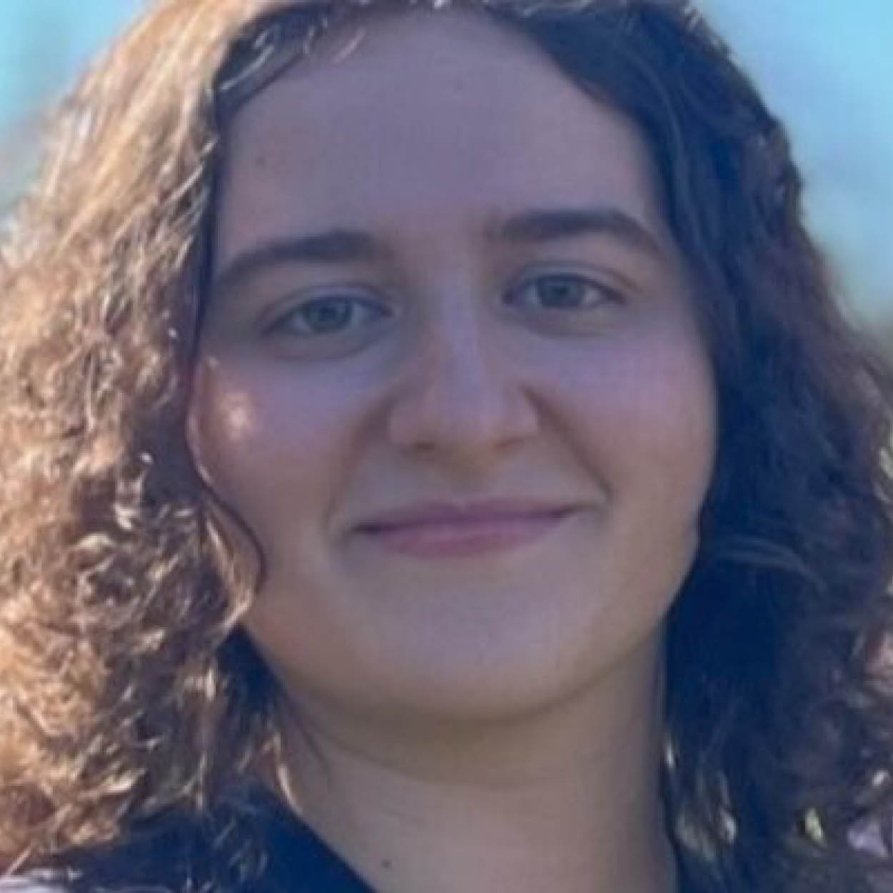 Close-up of a smiling woman with long, curly hair, wearing a dark top, outdoors with a cloudy sky in the background.