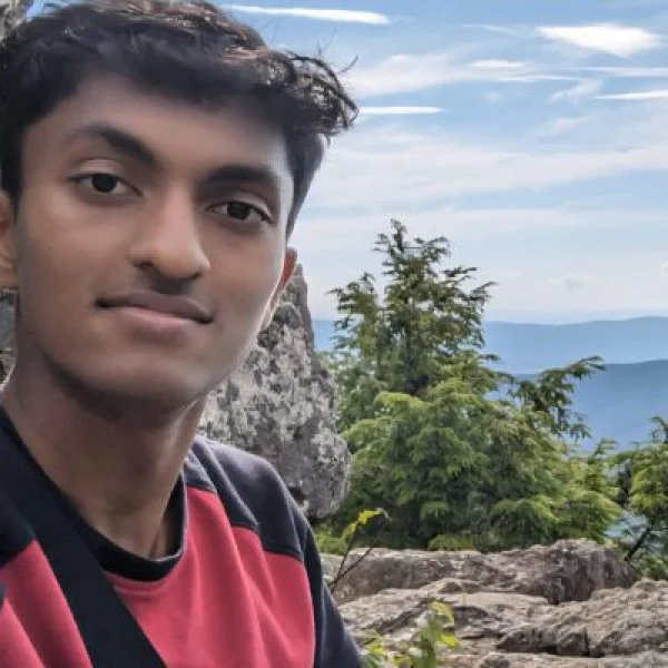 Young man with dark hair and medium skin tone taking a selfie outdoors on a rocky mountain with trees and a view of the sky and distant mountains in the background.