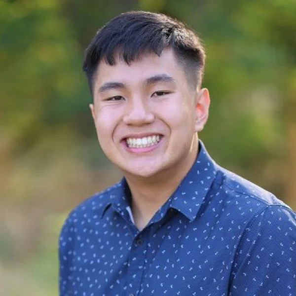 Young man with short dark hair smiling outdoors, wearing a blue button-up shirt with small white polka dots, blurred trees in the background.