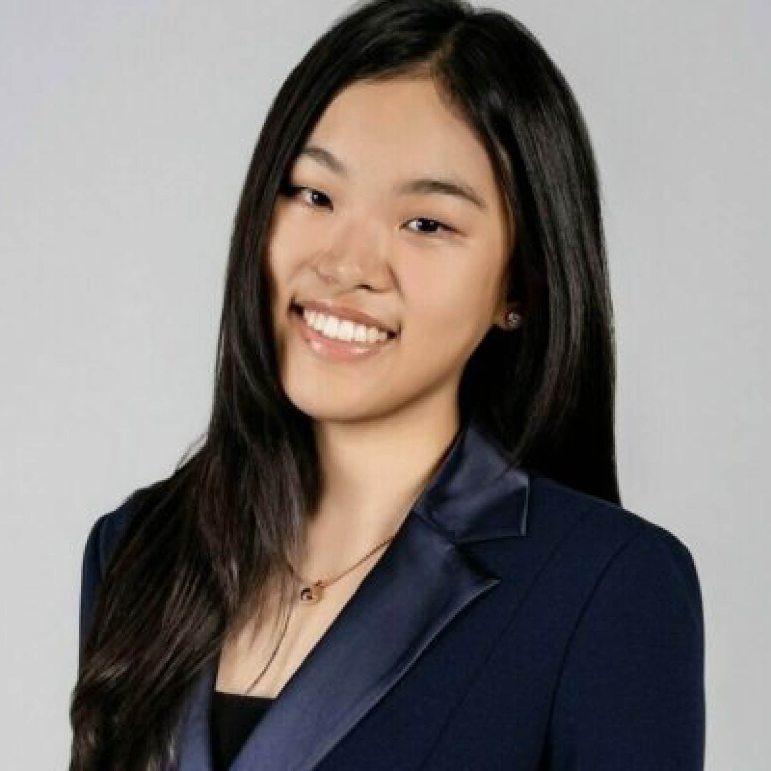 Woman with long dark hair, wearing a dark blazer and a necklace, against a plain light gray background.