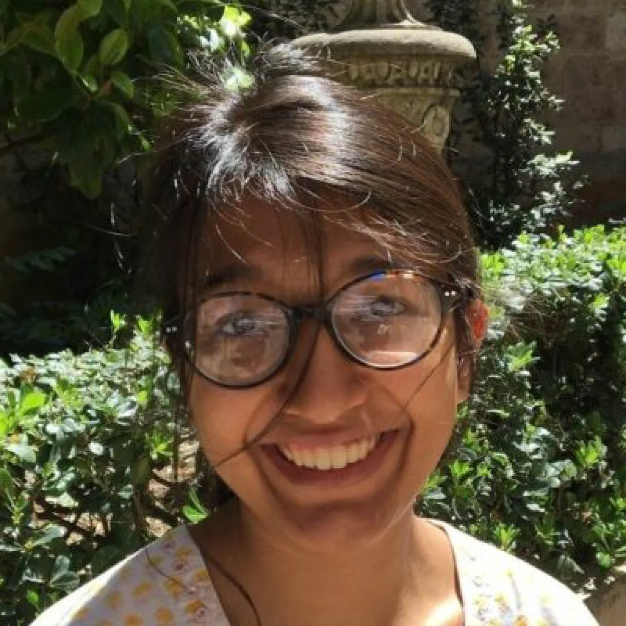 Smiling woman with glasses and short brown hair, outdoors surrounded by greenery and sunlight.