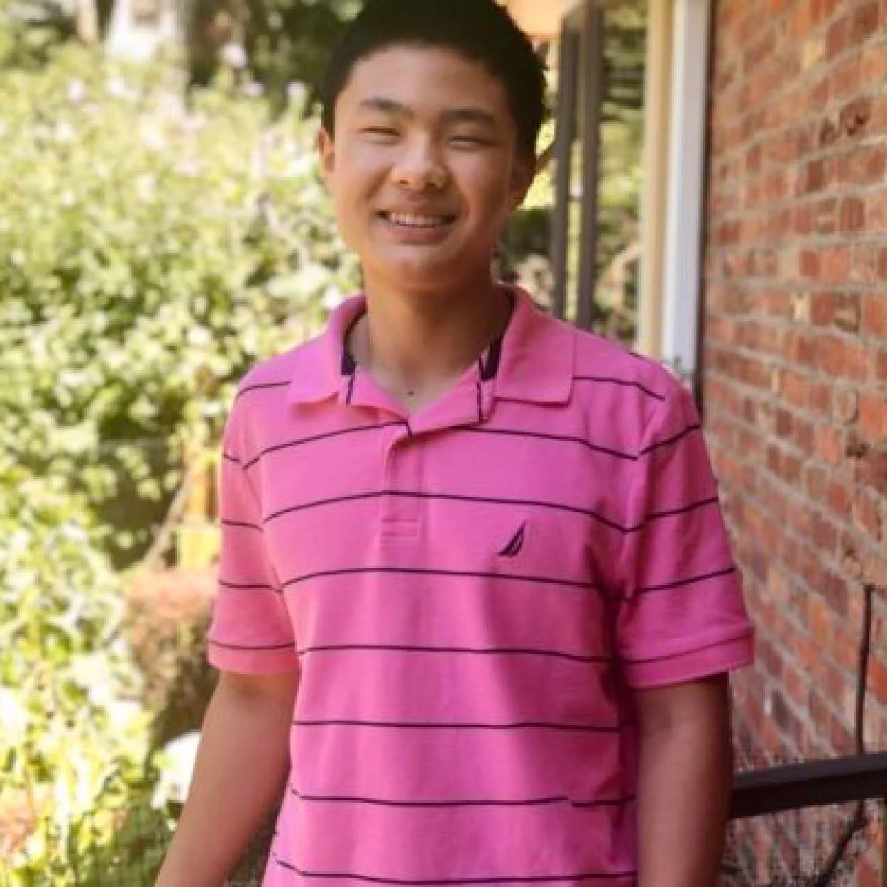 Young man smiling outdoors, wearing a pink striped polo shirt, standing next to a brick wall with trees and greenery in the background.