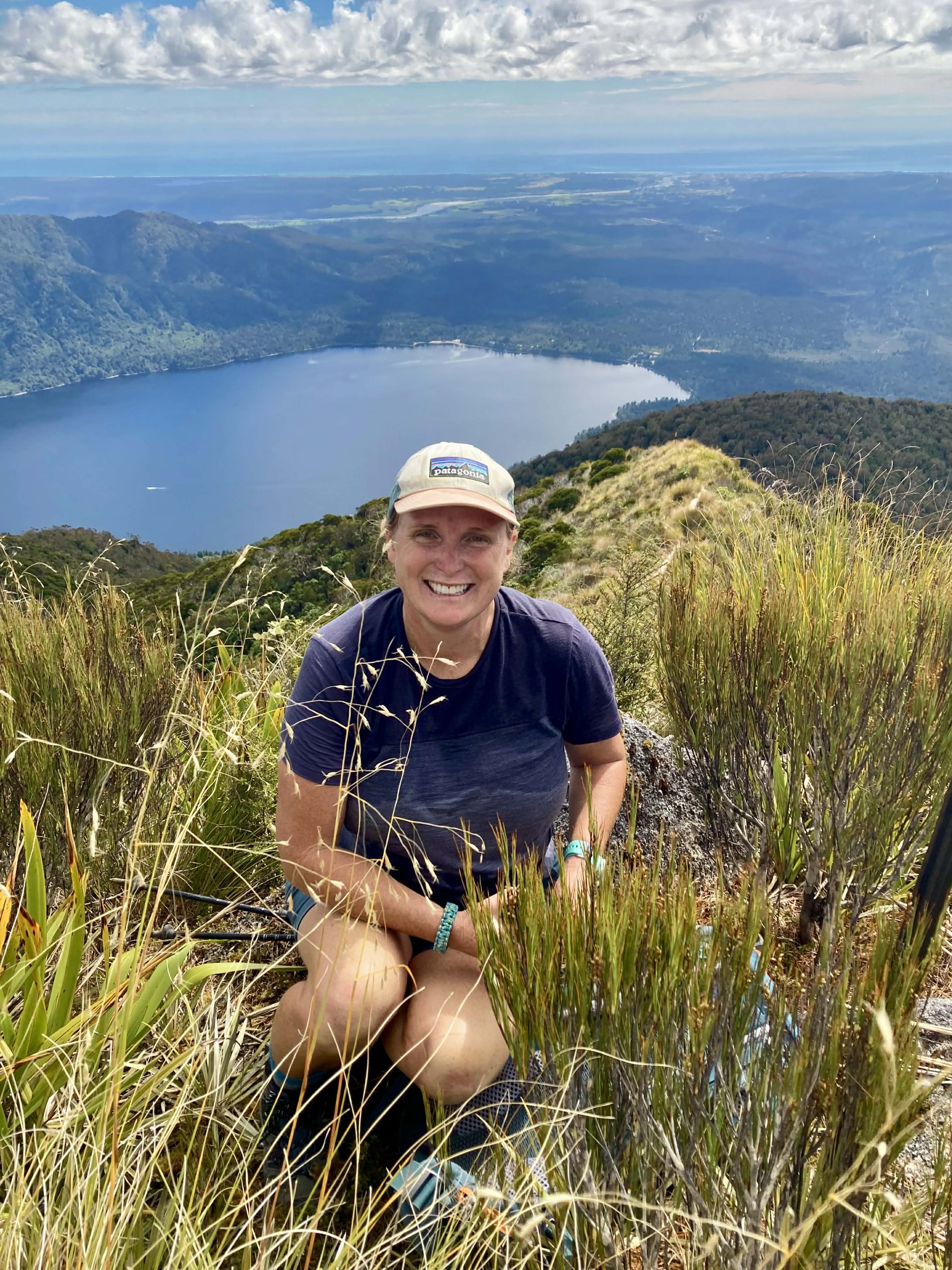 A smiling woman sitting on a grassy hillside overlooking a lake surrounded by green hills and mountains under a partly cloudy sky.
