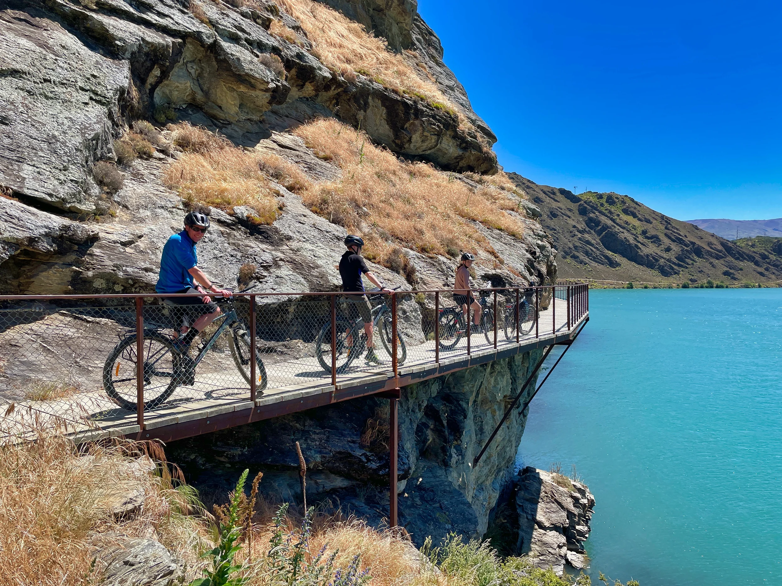People biking on a mountain trail with a rocky cliffside on the left and a calm blue lake on the right under a clear blue sky.