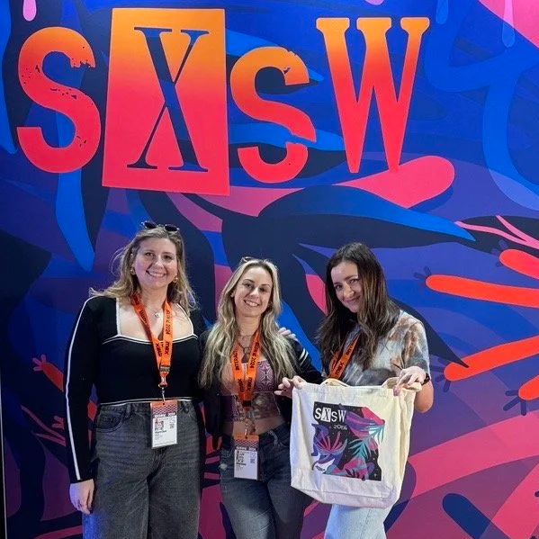 Three young women standing in front of a colorful SXSW sign, holding a tote bag with the SXSW logo.