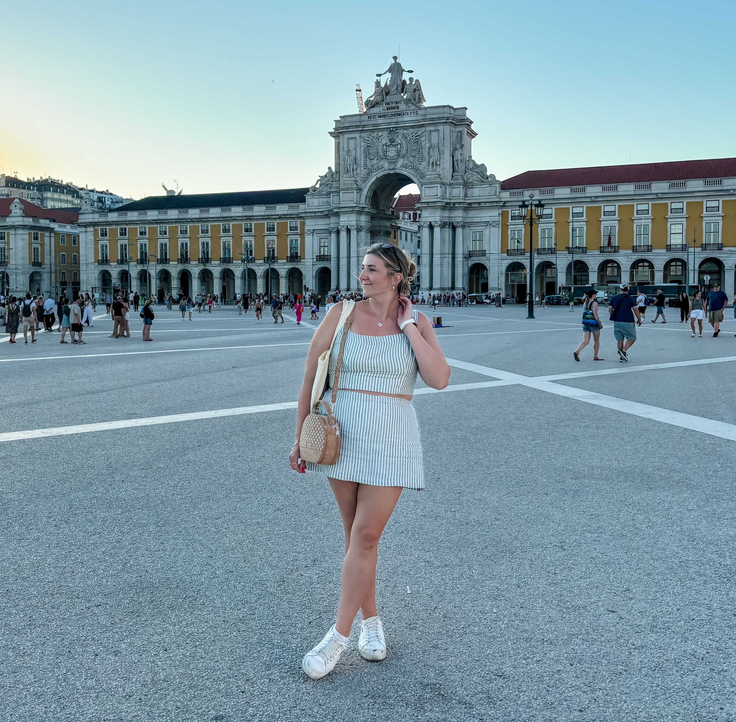 A young woman standing in a large public square with a historic archway adorned with sculptures behind her, and a crowd of people walking around.