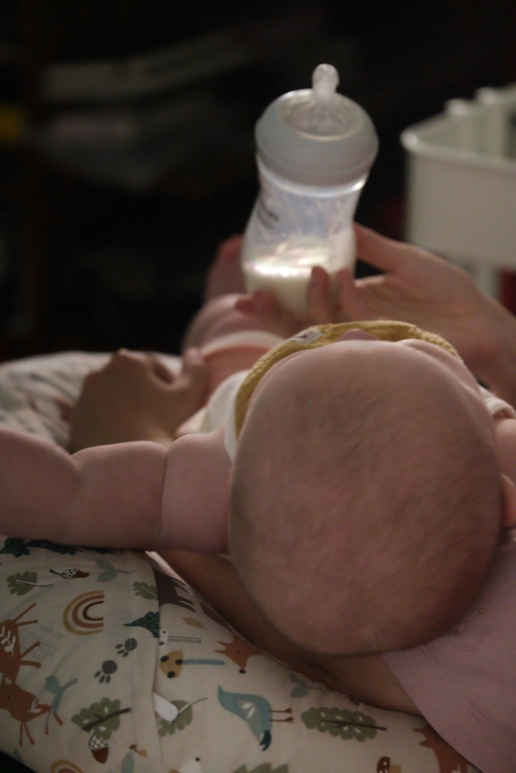 Close-up of a baby lying on a colorful blanket, drinking milk from a bottle held by an adult.