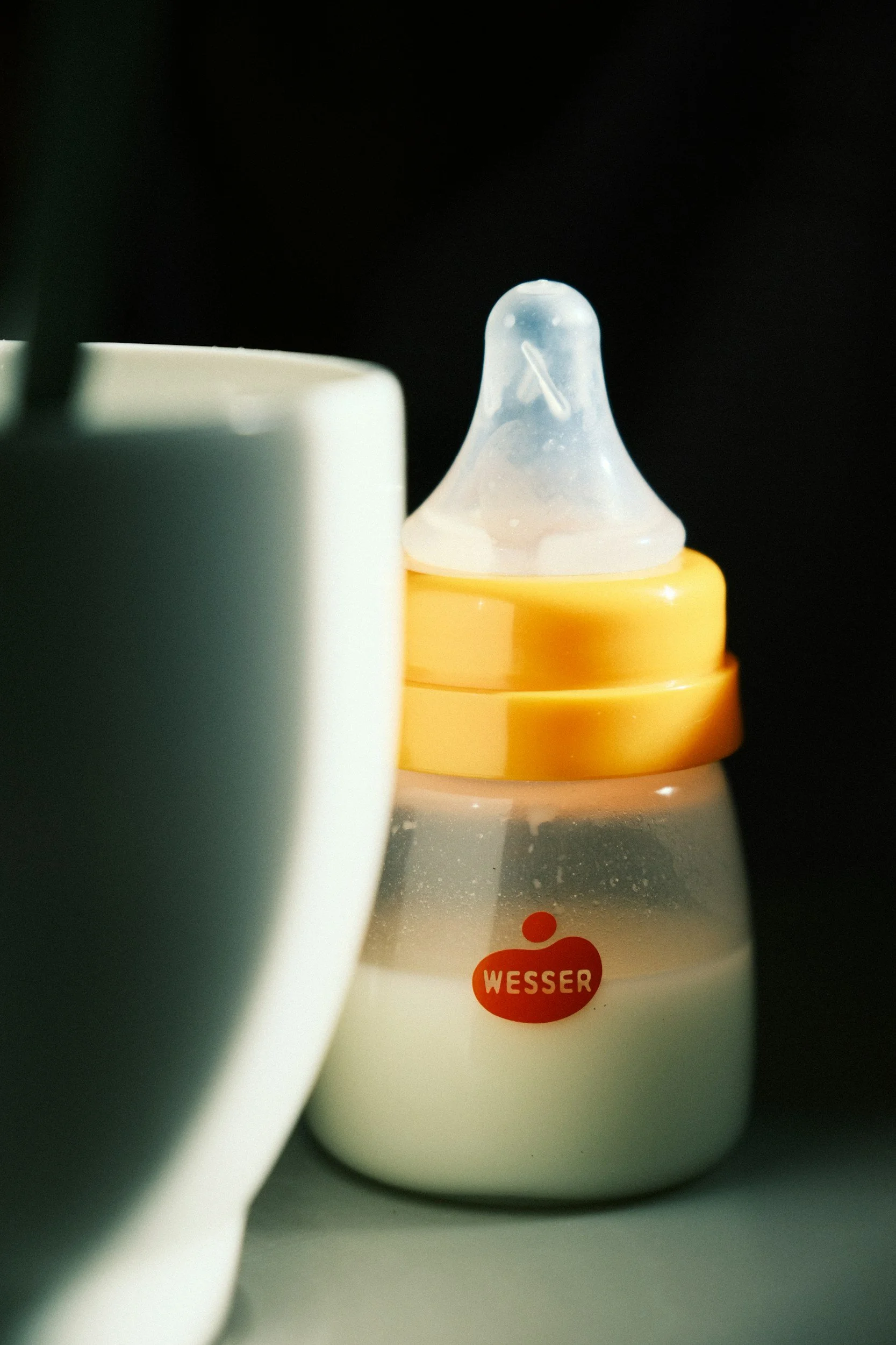 Close-up of baby bottle with Wesser logo, partially obscured by a white mug, featuring a silicone nipple on top.