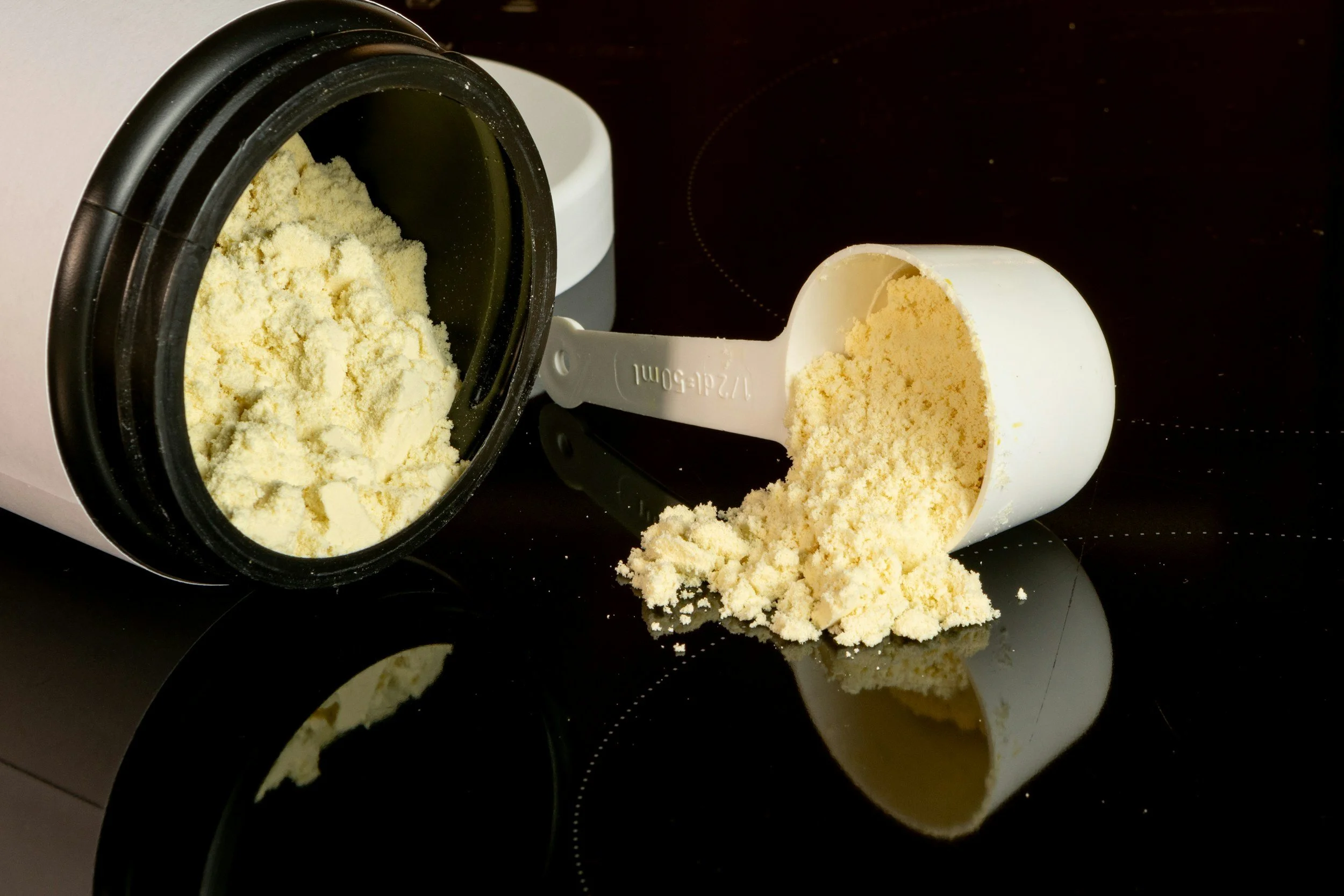 A black container with yellow powder, a measuring scoop filled with yellow powder, and some powder spilled on a black surface.