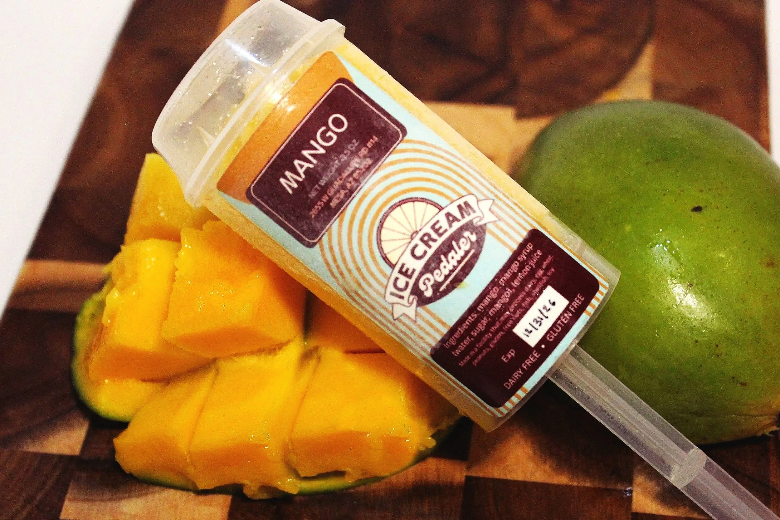 Close-up of a frozen mango push pop with a green mango and cut mango pieces on a wooden surface.