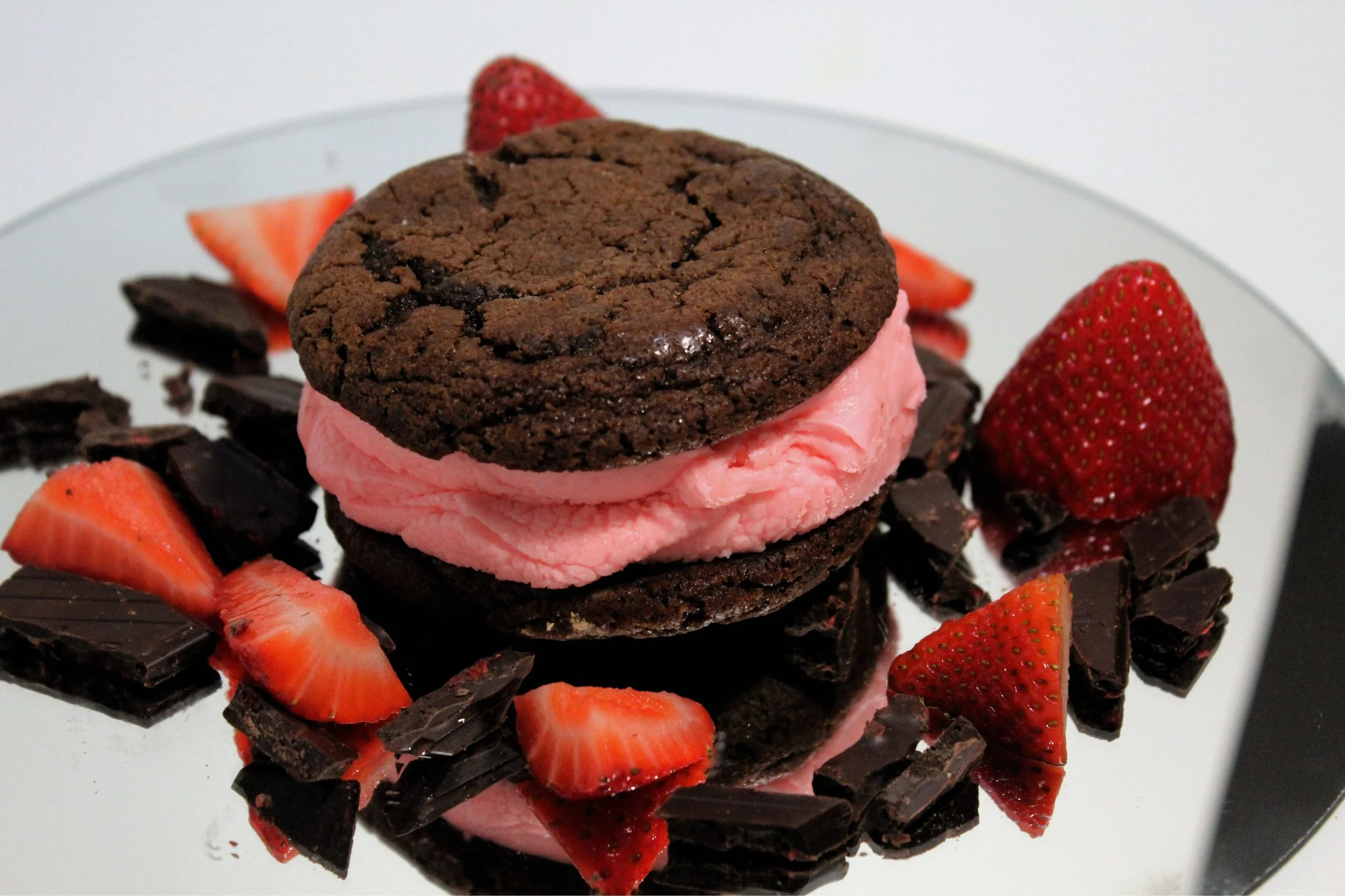 Chocolate chocolate chip cookies with strawberry ice cream in between, surrounded by sliced strawberries and pieces of dark chocolate on a white reflective surface.