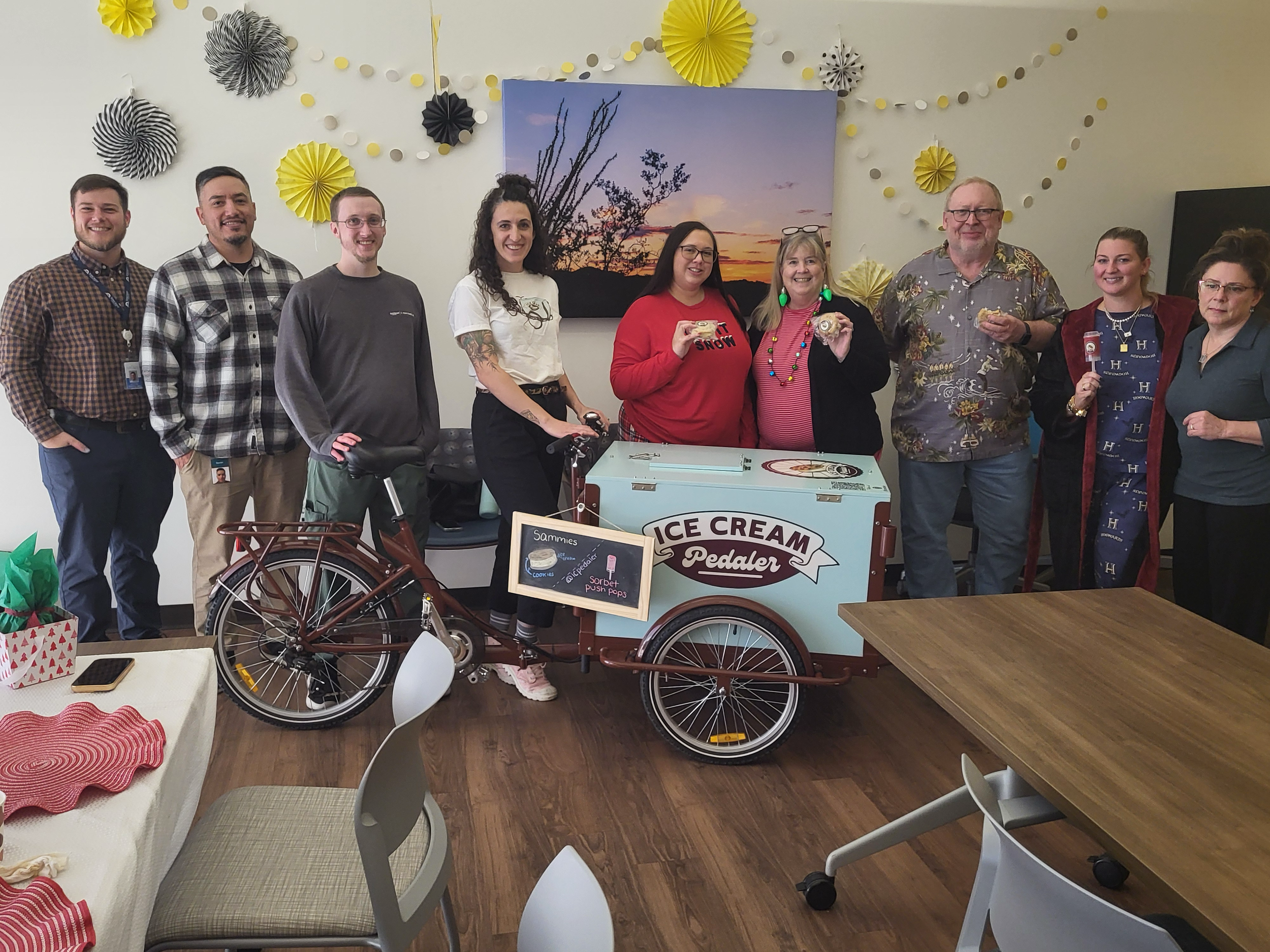 Group of nine people standing behind an ice cream cart decorated for a celebration in an indoor setting, with paper decorations on the wall, and tables in the foreground.