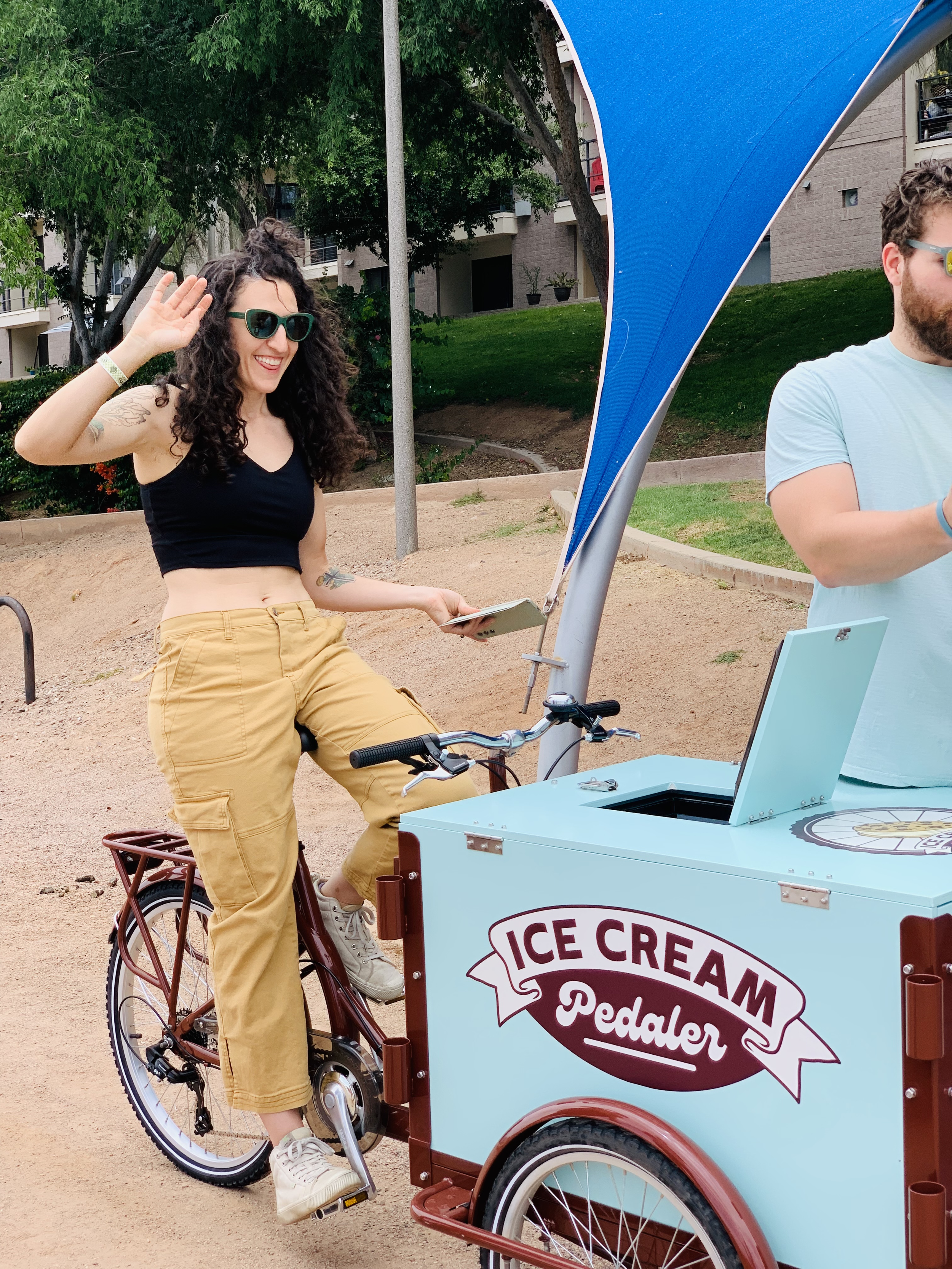 A woman with curly hair, wearing sunglasses, a black crop top, and beige pants, riding a tricycle ice cream cart, waving and smiling