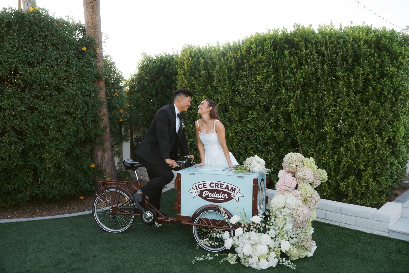 A bride and groom dressed in wedding attire are leaning towards each other and smiling, standing behind a vintage ice cream bike cart decorated with white flowers, in a garden with green hedges and trees.
