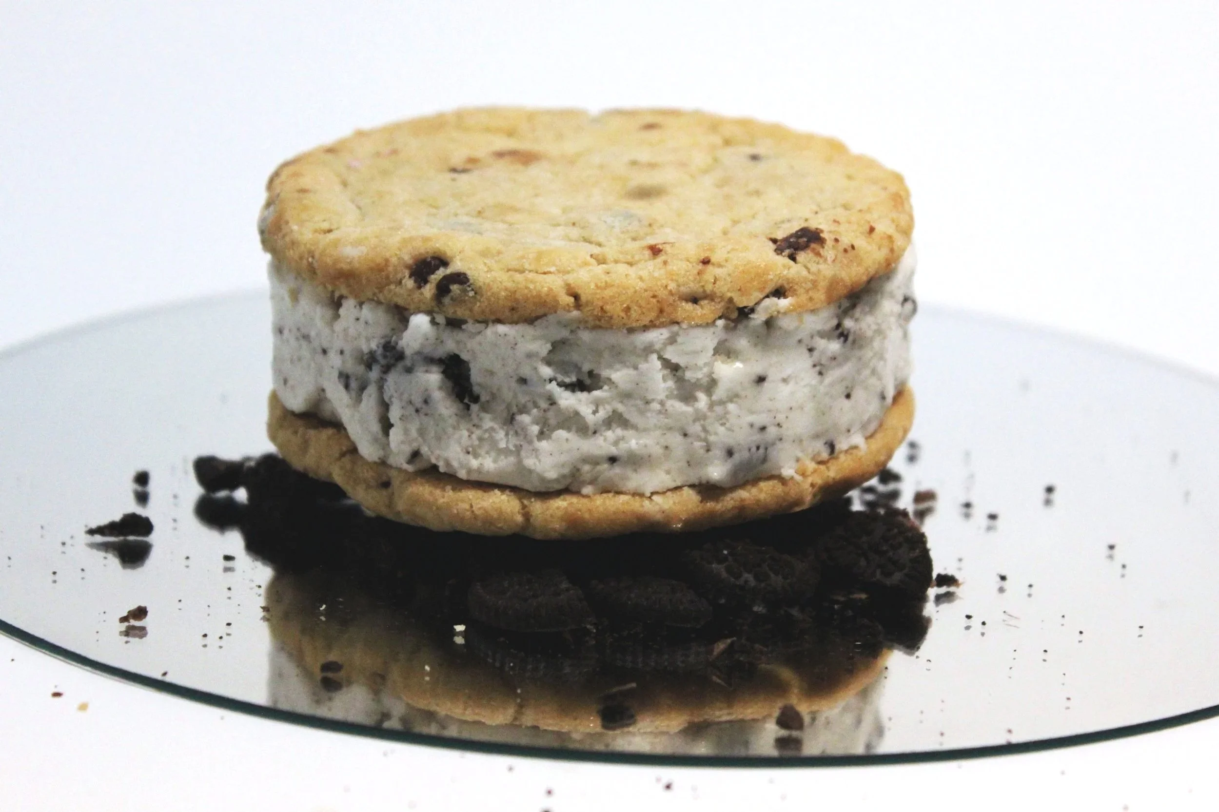 A cookies and cream ice cream sandwich with chocolate chip cookies, placed on a reflective surface with sandwich cookie crumbs