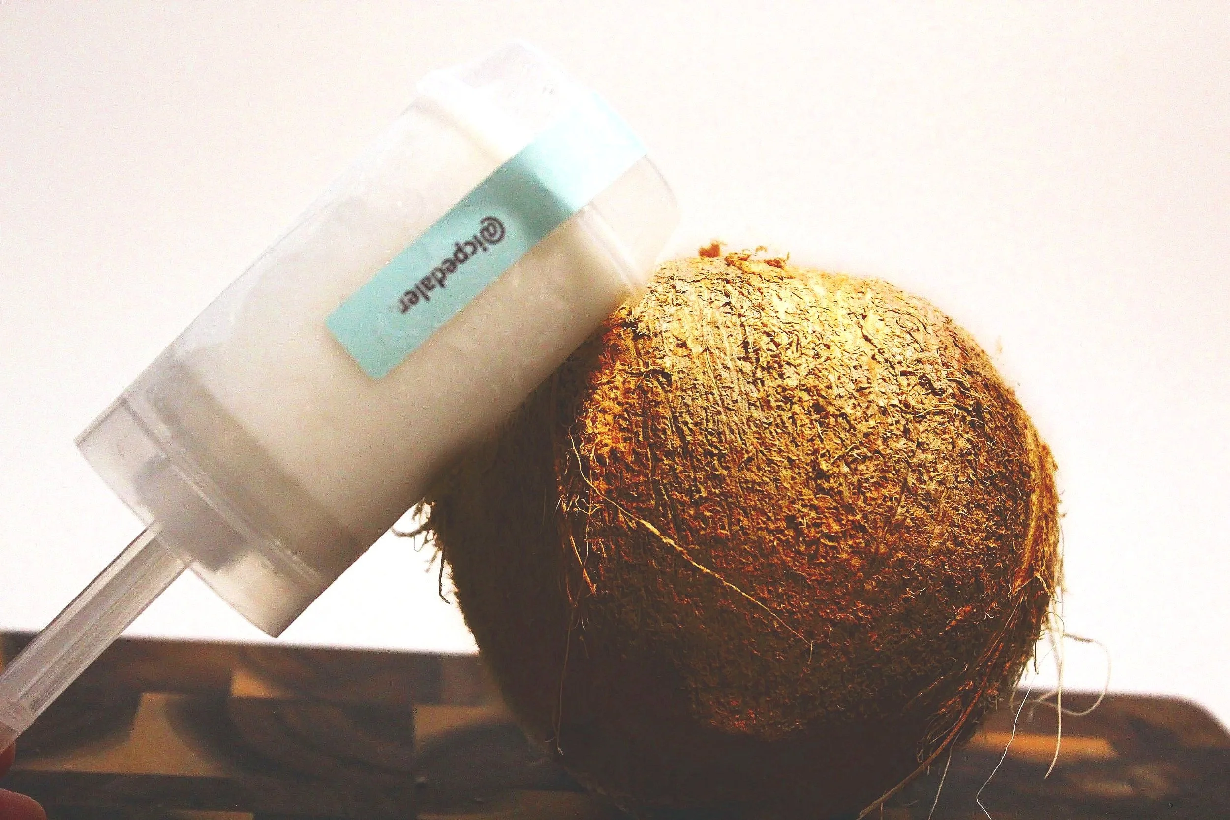 A coconut with rough brown outer shell next to a coconut push pop against a plain white background.