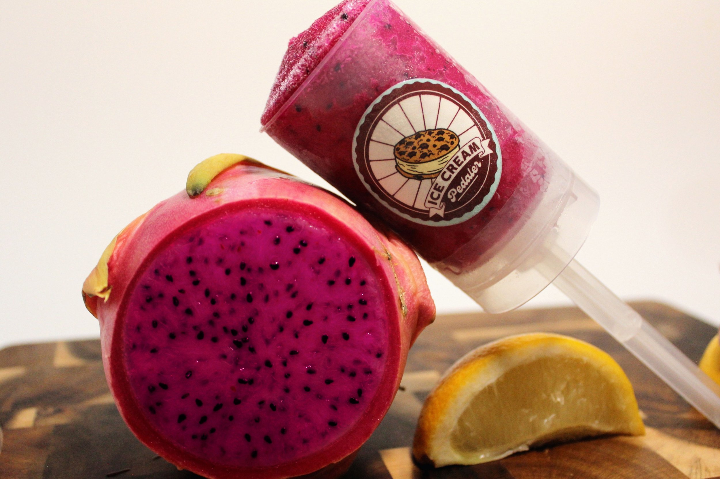 Close-up of a dragon fruit sliced in half, showing its vibrant pink flesh with black seeds, along with slices of lemons in the background with a dragon fruit sorbet push pop